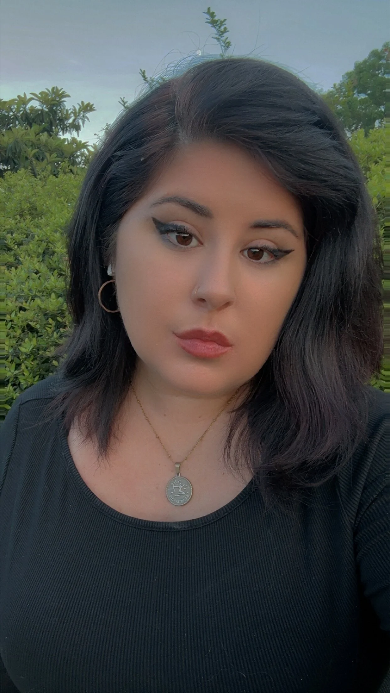 Close-up photo of a young woman with black hair, brown eyes, and makeup including winged eyeliner, wearing a black top, a gold necklace, and hoop earrings, standing outdoors with greenery and trees in the background.