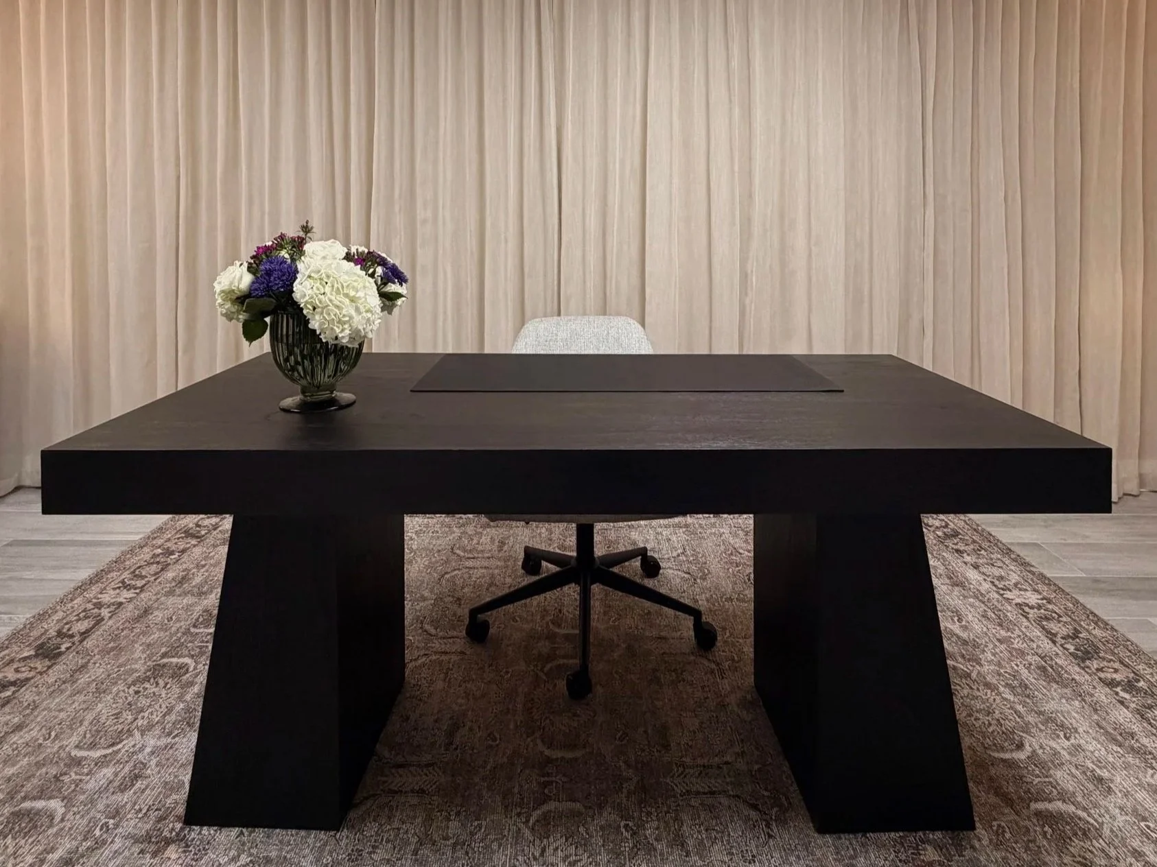 Modern office meeting room with a large black table, a gray office chair, a vase of white and purple flowers on the table, beige curtains in the background, and a patterned rug underneath.