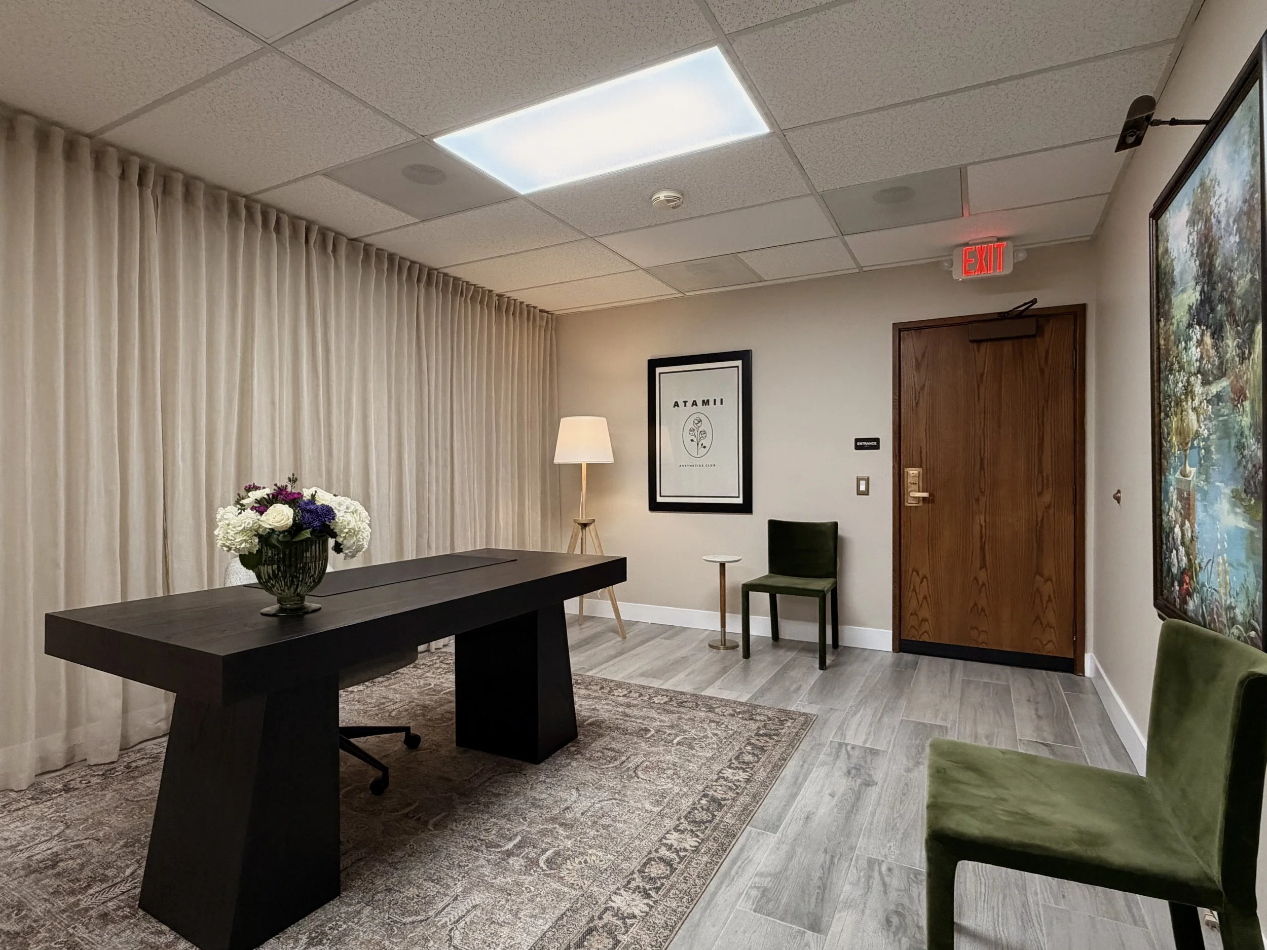 The Atamii lobby with a dark wooden desk, a vase of white and purple flowers, a beige rug, green and black chairs, a tall lamp, framed artwork, cream-colored curtains, and a closed wooden door with an exit sign above.