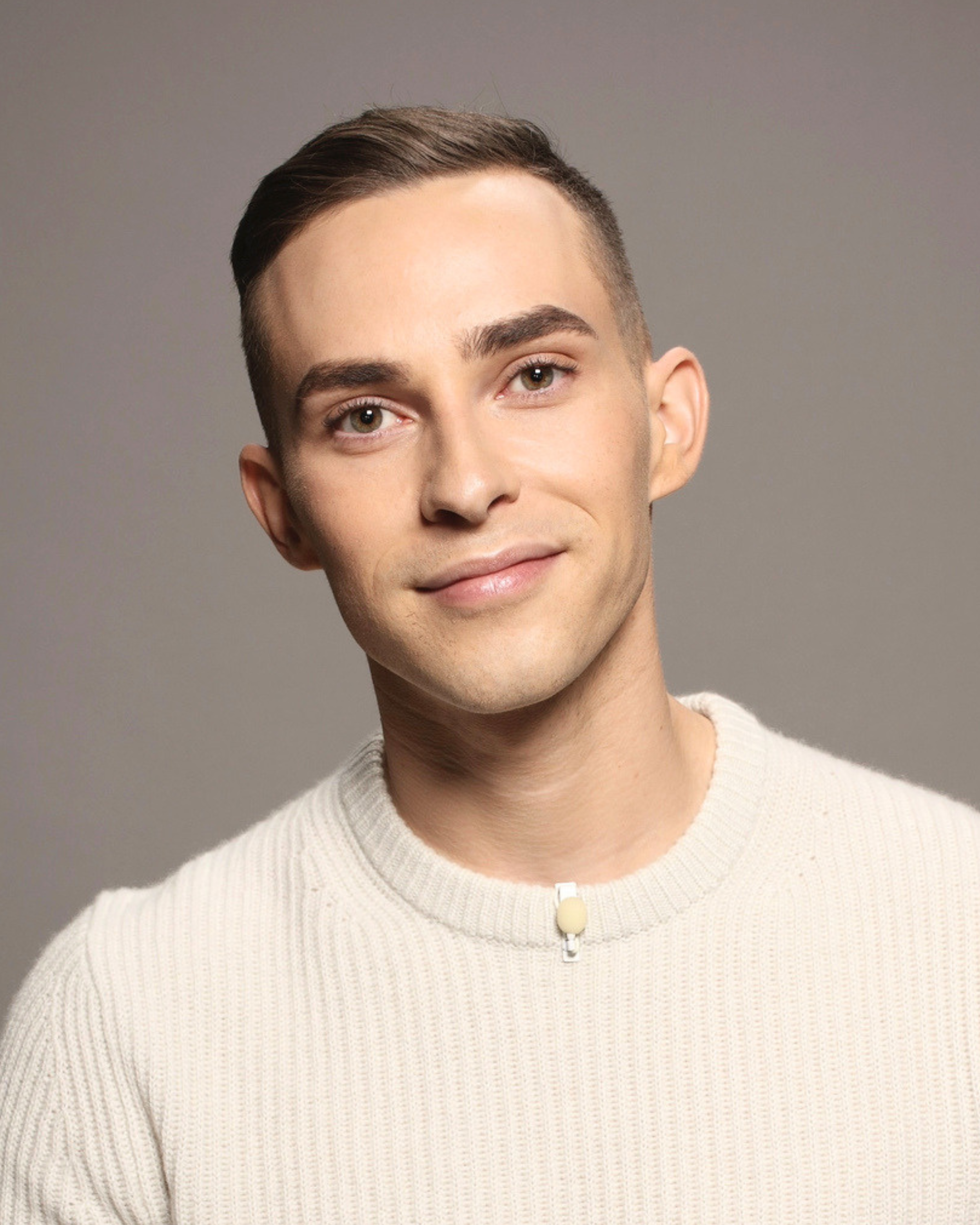 Portrait of a young man with short brown hair, fair skin, and wearing a cream-colored sweater, standing against a plain gray background.