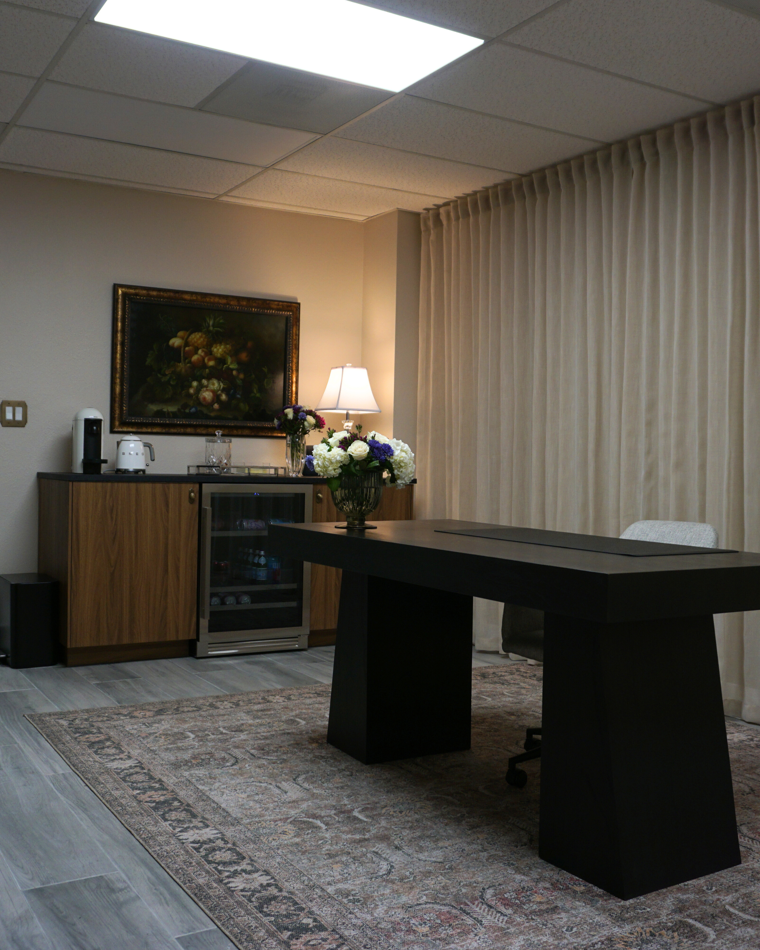 A hotel conference room with beige curtains, a large black table, a floral centerpiece, and a wooden cabinet with a coffee maker, a lamp, and a framed painting of fruit.