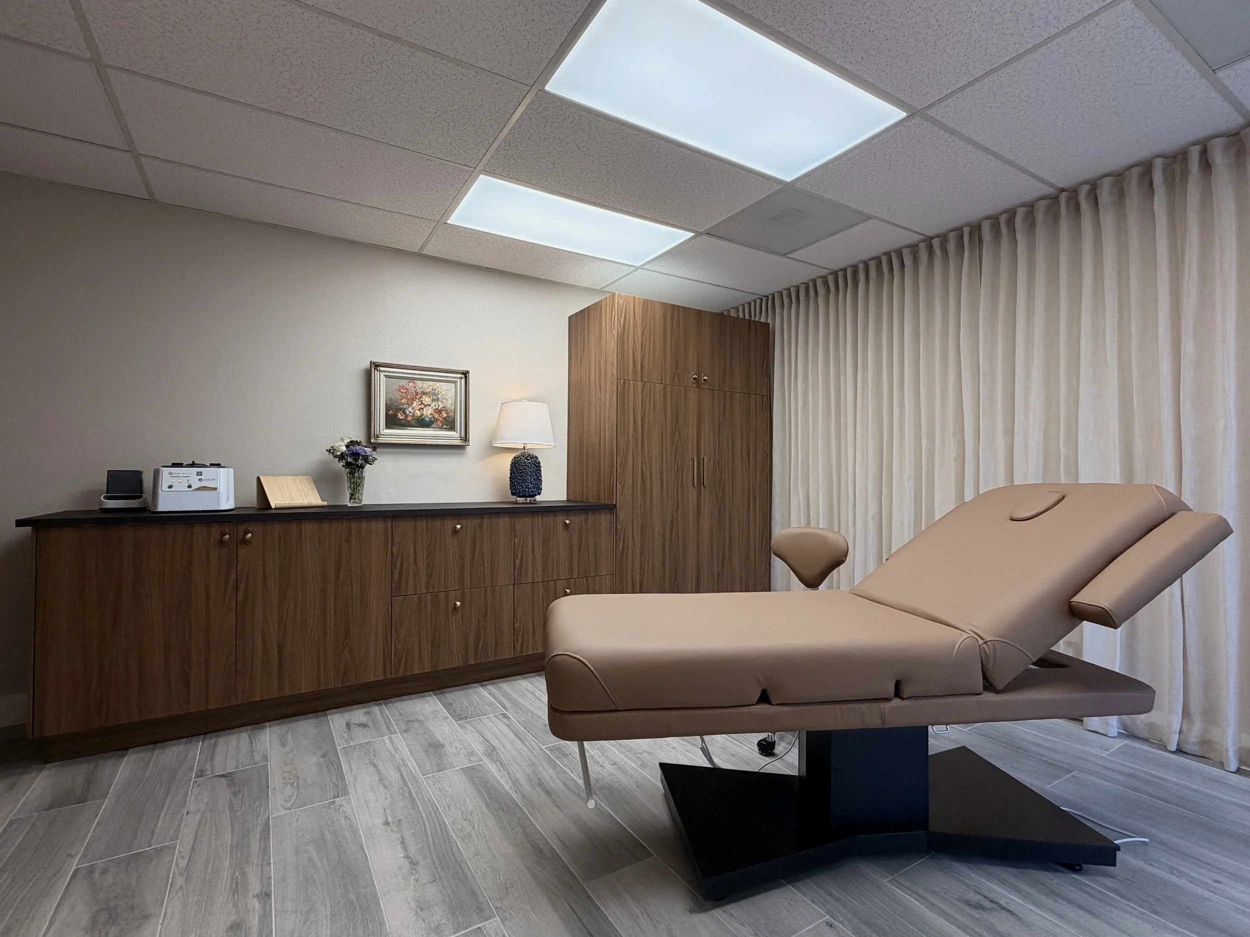 A medical examination room with a beige reclining examination table, a wooden cabinet, a desk with a painting, a lamp, a small vase with flowers, and curtains covering a window.