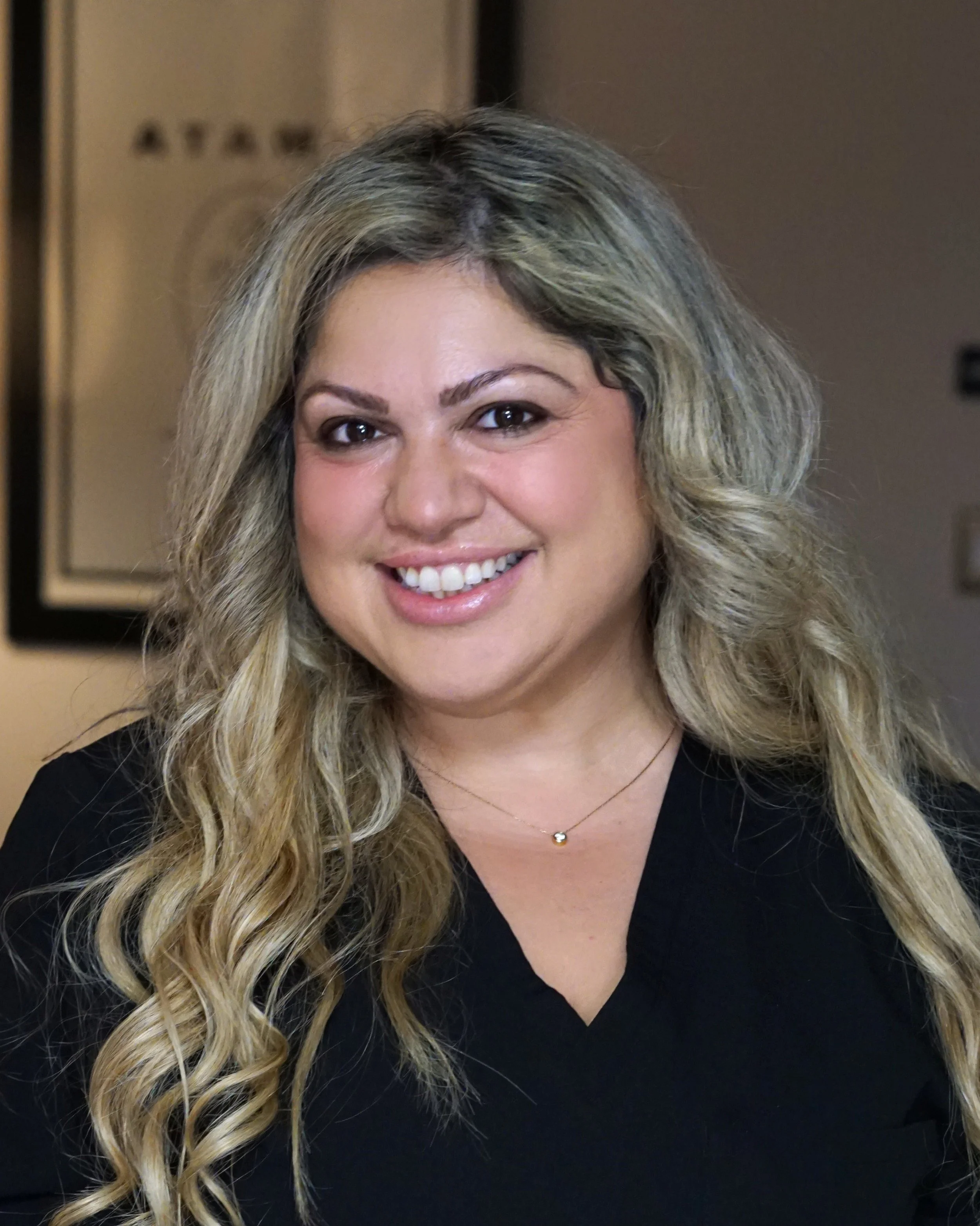A woman with long blonde curly hair, smiling, wearing a black top and a delicate necklace.