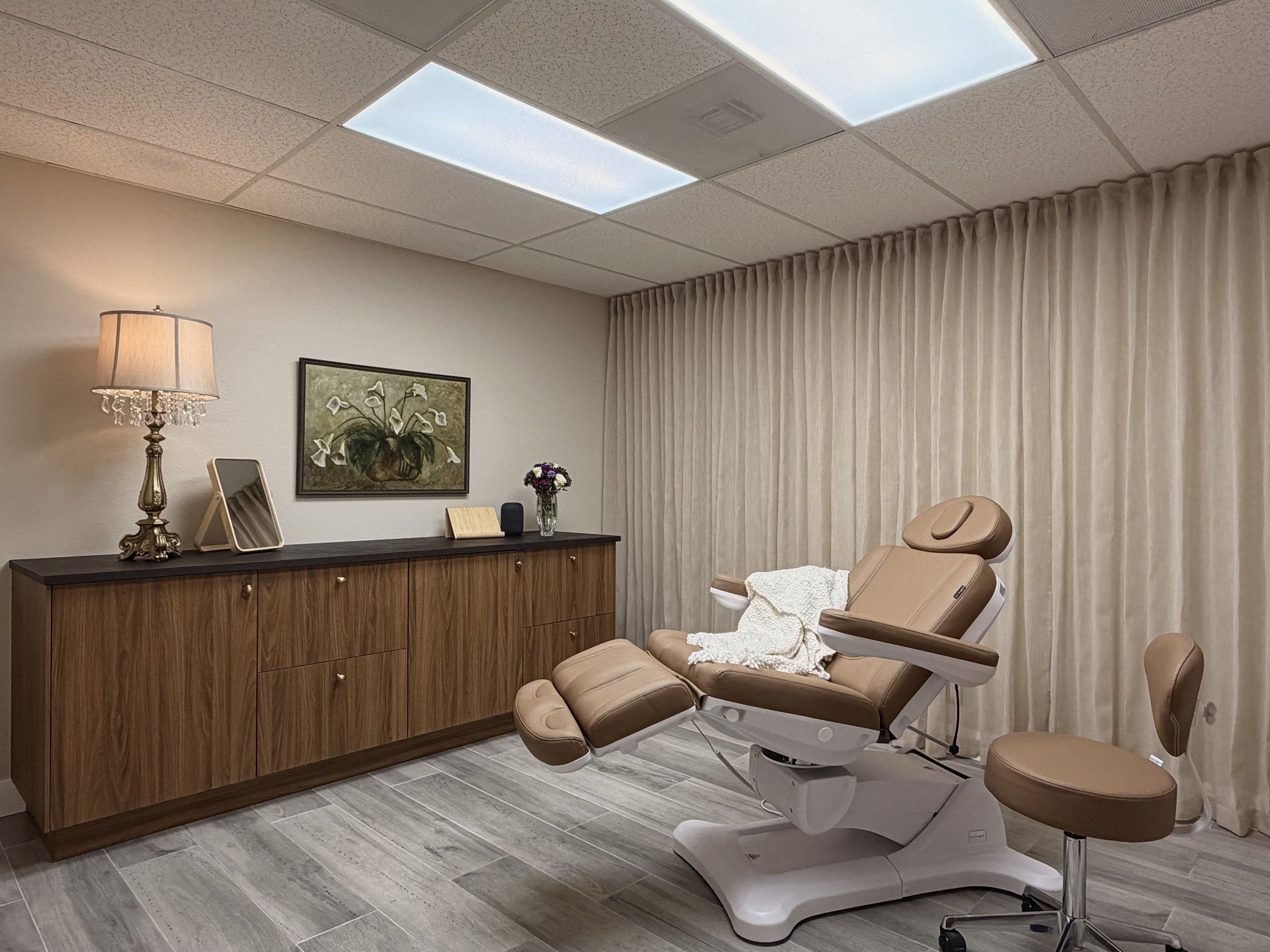 Medical examination room with an adjustable beige treatment chair, a wooden cabinet with decorative items, and beige curtains, lit by ceiling lights and a table lamp.
