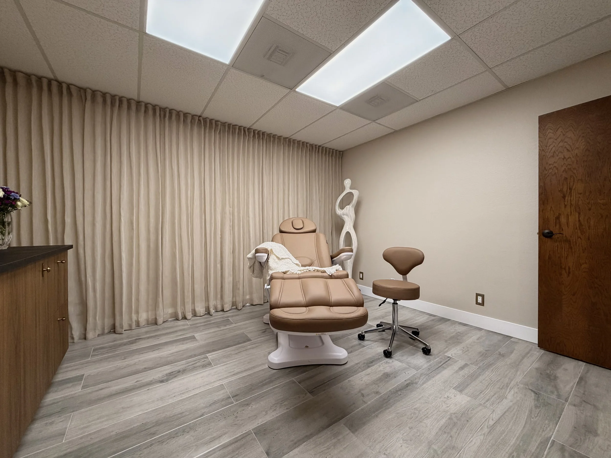 A medical examination room with a beige examination chair, a beige rolling stool, a decorative white sculpture, beige curtains, and a wooden cabinet on the left.
