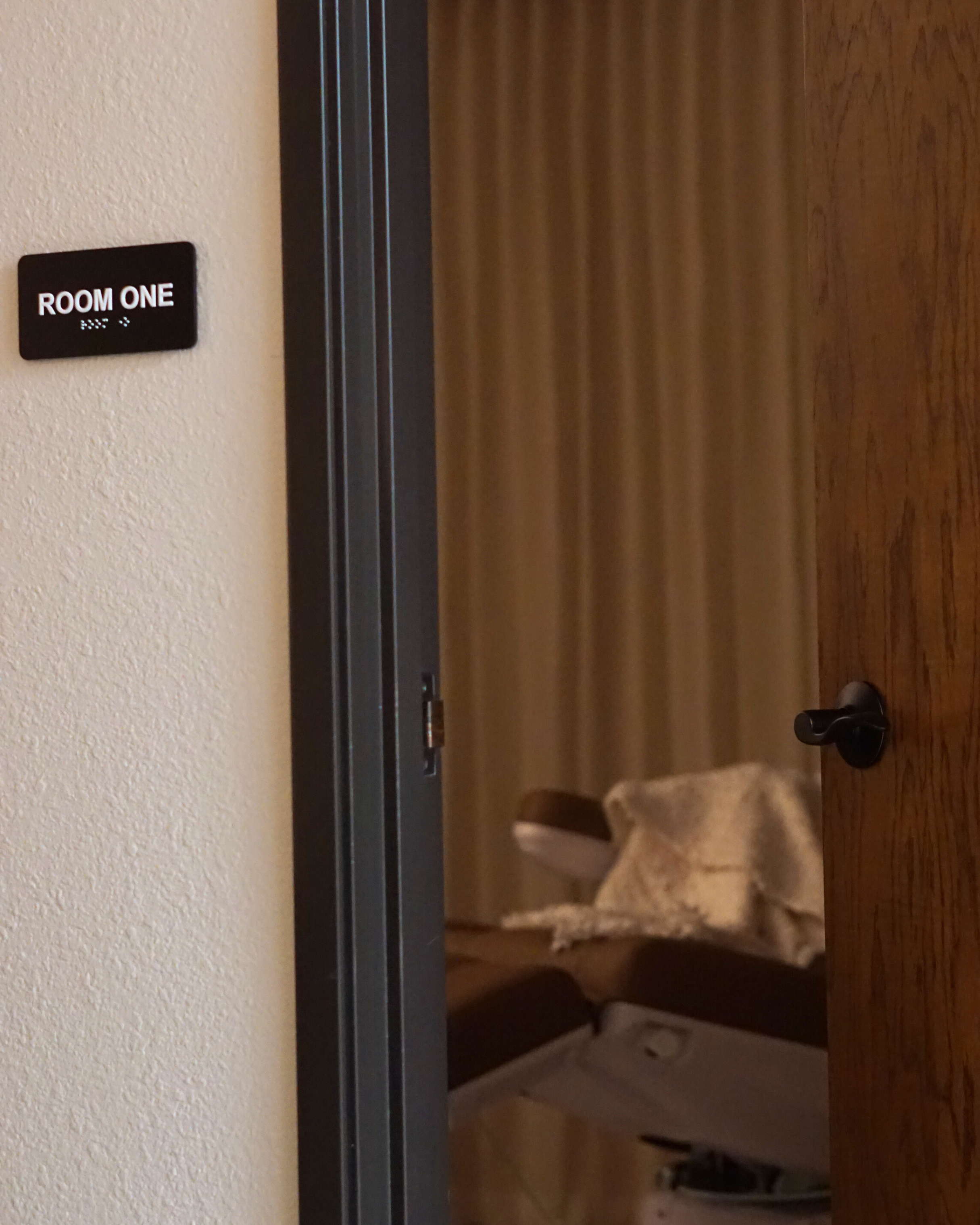 A partially open wooden door revealing a treatment room with a padded table, pillow, and beige curtains, and a small sign on the wall reads 'ROOM ONE' with Braille underneath.