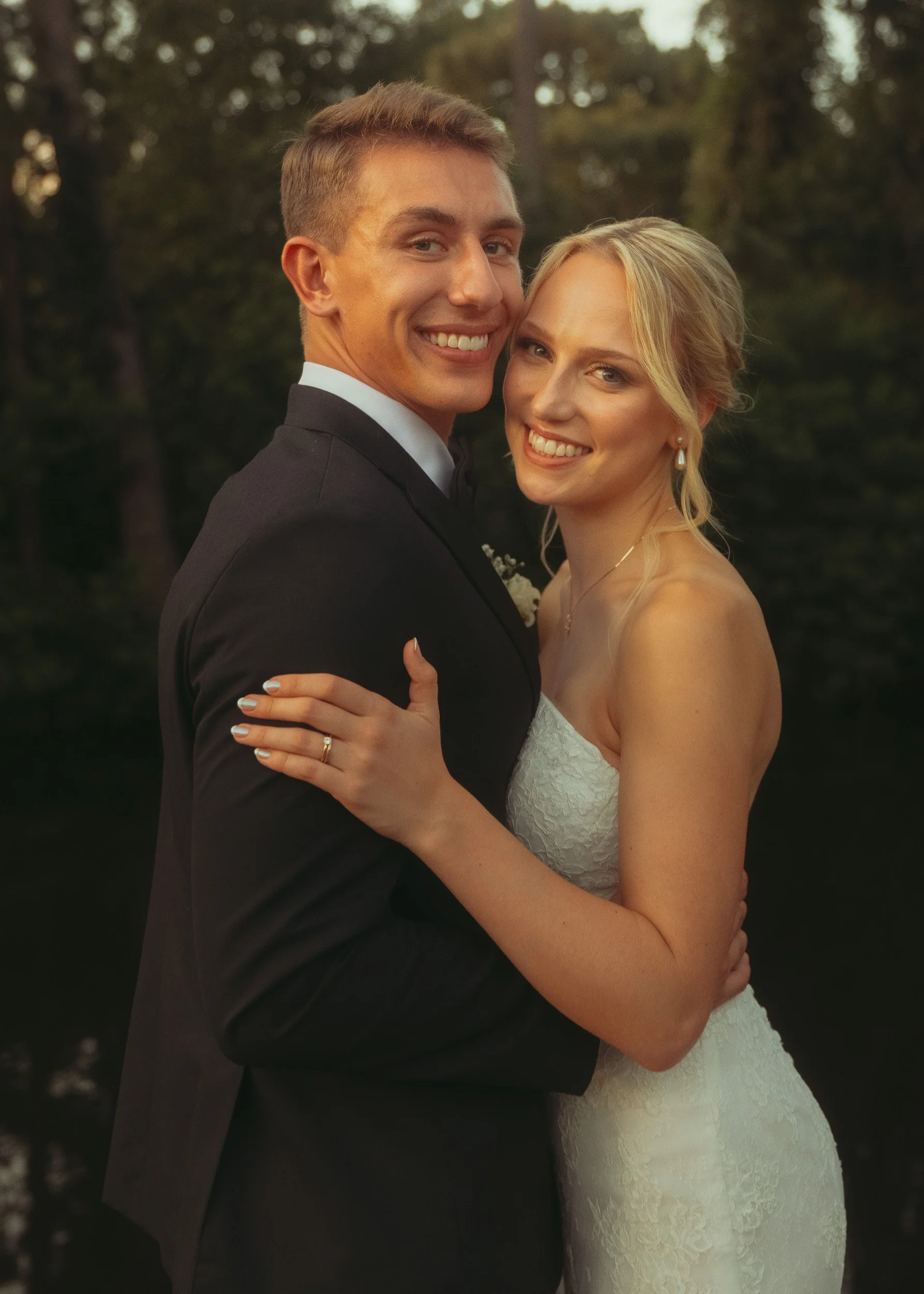 A newlywed couple smiling and hugging outdoors during sunset, the groom in a black tuxedo and the bride in a white lace strapless wedding dress.