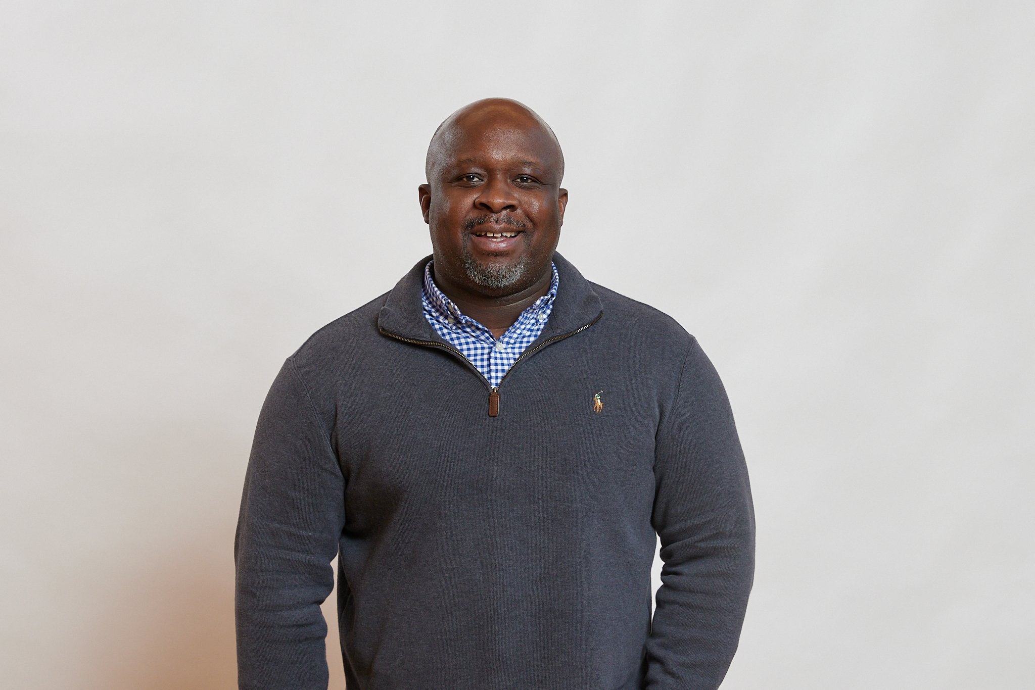 A man with dark skin, a shaved head, and a beard, wearing a dark gray pullover and a blue checkered shirt, standing against a plain white background, smiling at the camera.