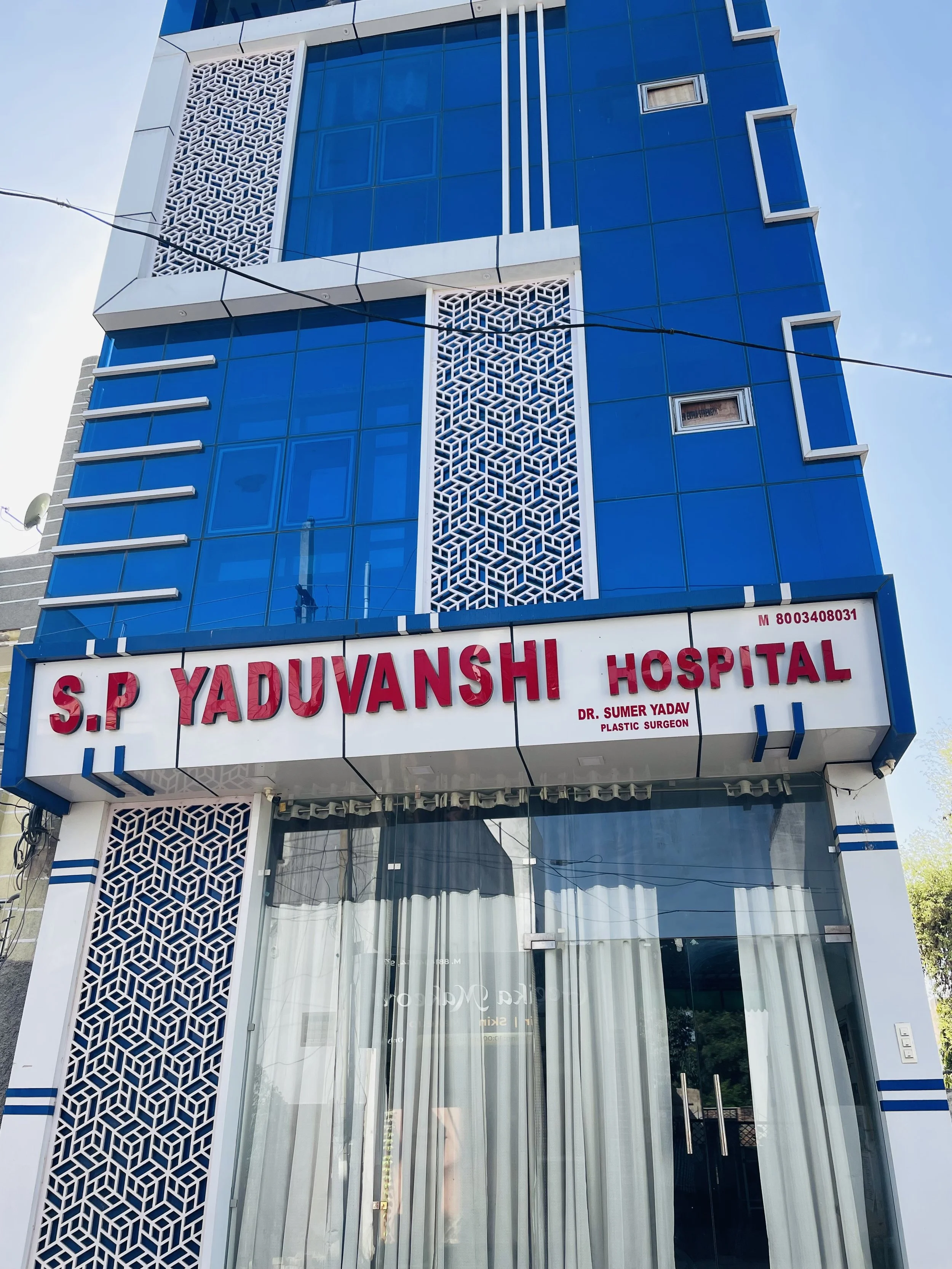 The image shows the front of a modern building with blue glass windows and decorative white lattice panels. There is a sign that reads "S.P. YADUVANSHI HOSPITAL" in red letters, along with the name of Dr. Sumer Yadav, a plastic surgeon. The building has vertical white and blue accents, and large glass doors with curtains behind them.