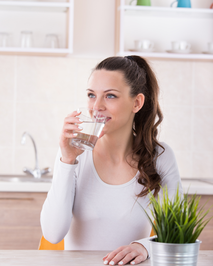 A woman with long brown hair in a ponytail drinking a glass of water in a bright kitchen with white walls and a potted plant on a table.