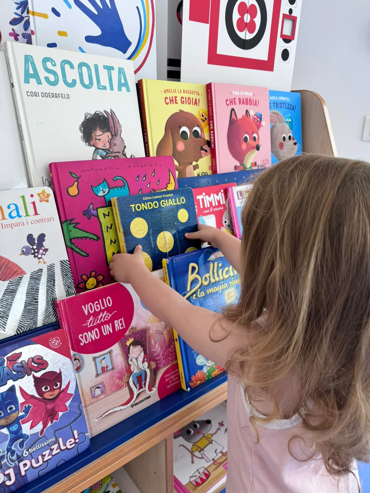 Un bambino con capelli castani riccioli cerca di prendere un libro rosa con una tigre sulla copertina; ci sono diversi libri colorati disposti ordinatamente su uno scaffale.