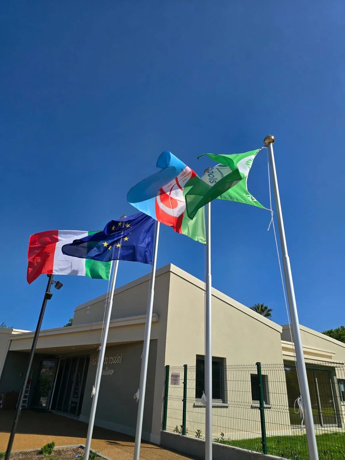 Bandiera dell'Unione Europea, Italia, e una bandiera verde, tutte issate davanti a un edificio moderno sotto un cielo blu limpido.