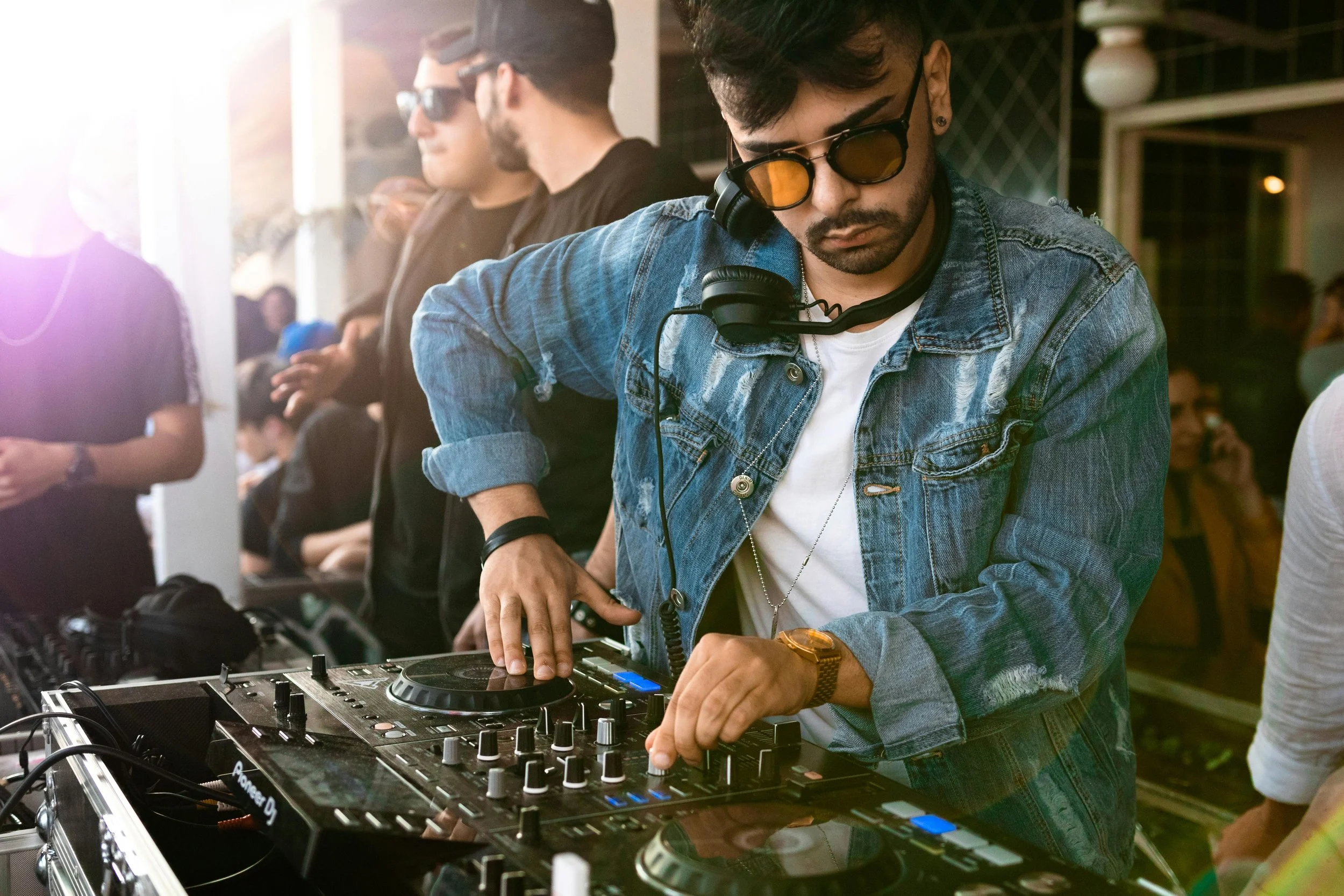 A DJ wearing sunglasses and a denim jacket is mixing music on a DJ controller at a party or nightclub, with people dancing in the background under warm lighting.