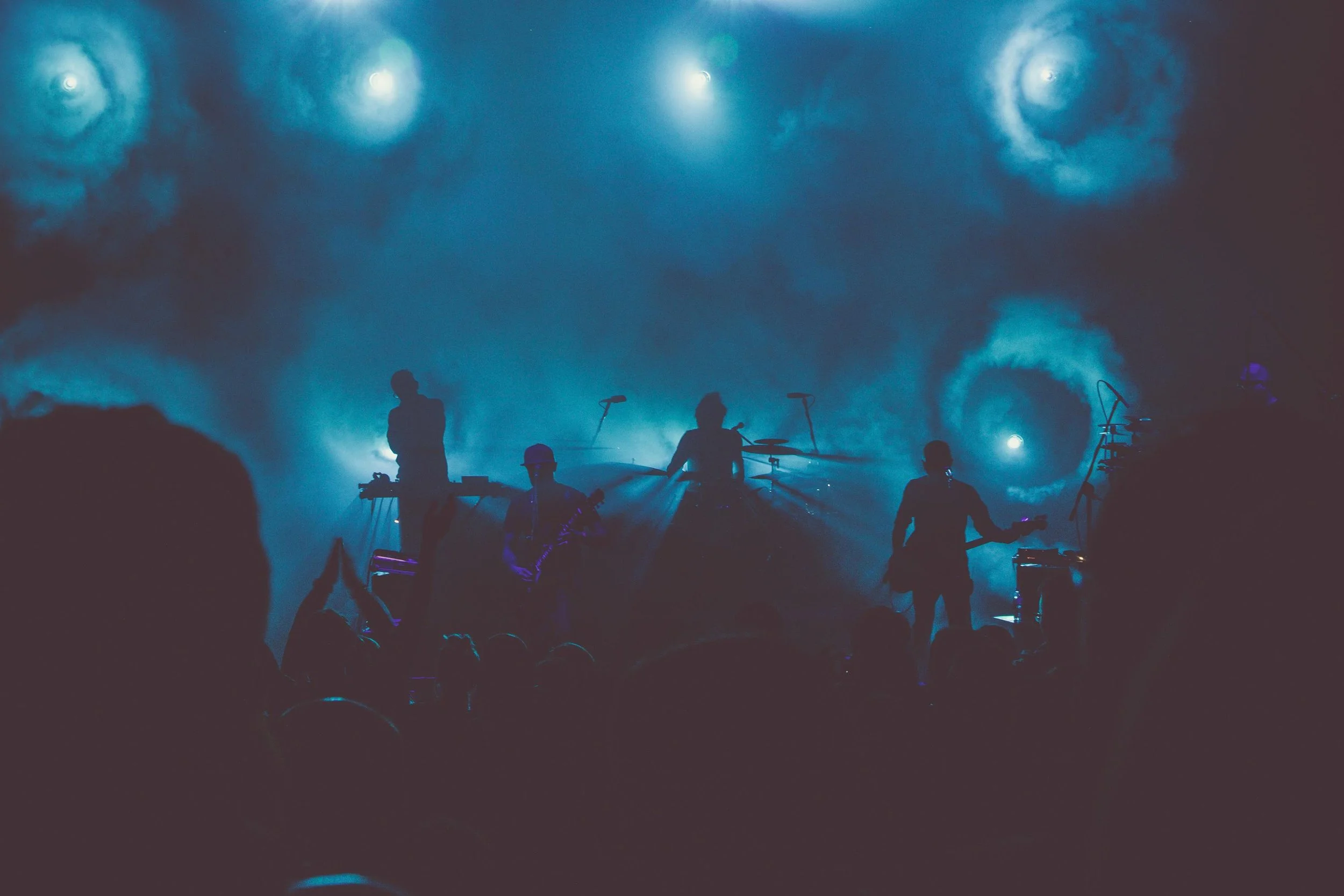 Silhouettes of a band performing on stage with blue lighting and fog effects, audience visible in foreground.