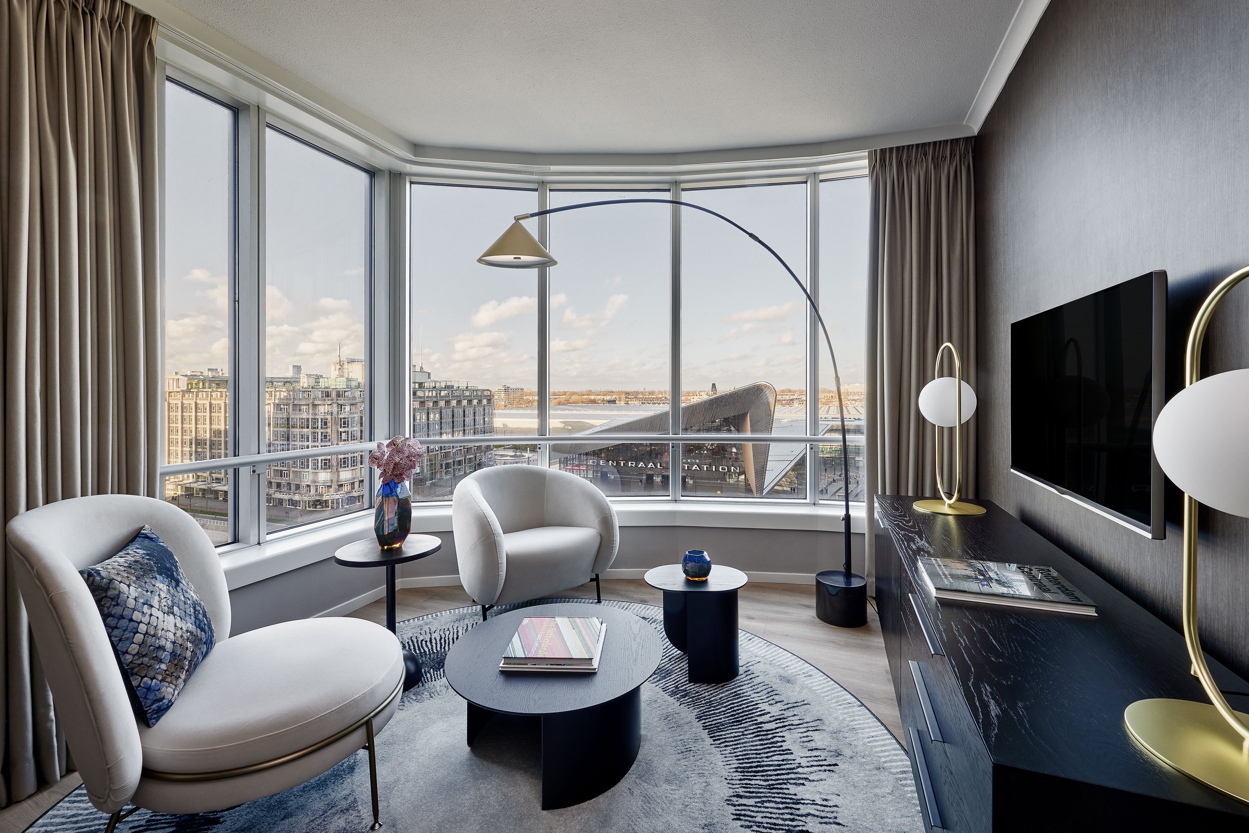 Marriott Rotterdam suite living area with curved white armchairs, round dark coffee table, gold pendant light, and panoramic city views through floor-to-ceiling windows