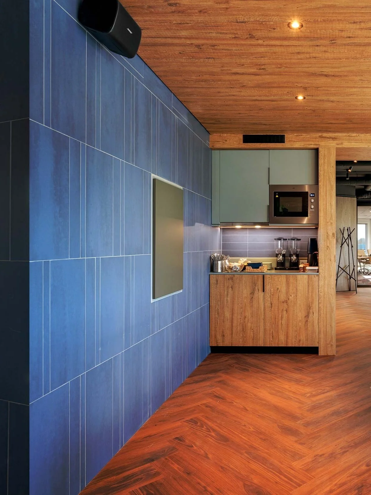 NSI Head Office Amsterdam corridor with blue ceramic tile feature wall, warm hardwood herringbone flooring, recessed ceiling lights, and glass-partitioned meeting room at the end