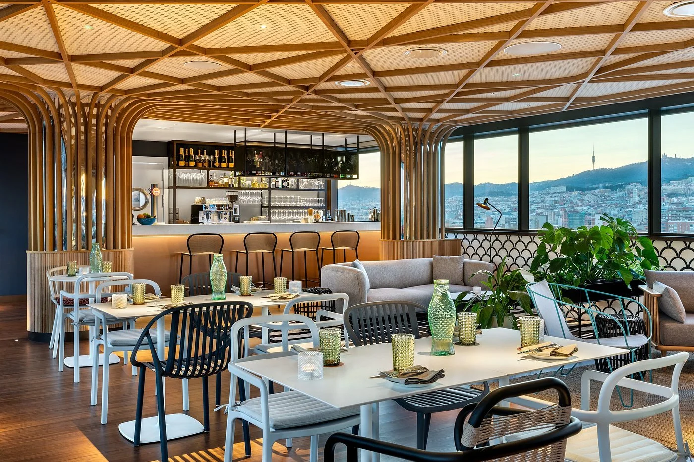 Modern restaurant interior with various tables and chairs, a bar area with barstools, large windows with city views, and contemporary decor including green vases and plants.