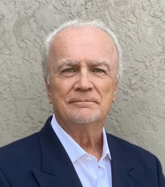 Portrait of an elderly man with gray hair and a goatee, wearing a dark suit and white shirt, standing in front of a textured wall.
