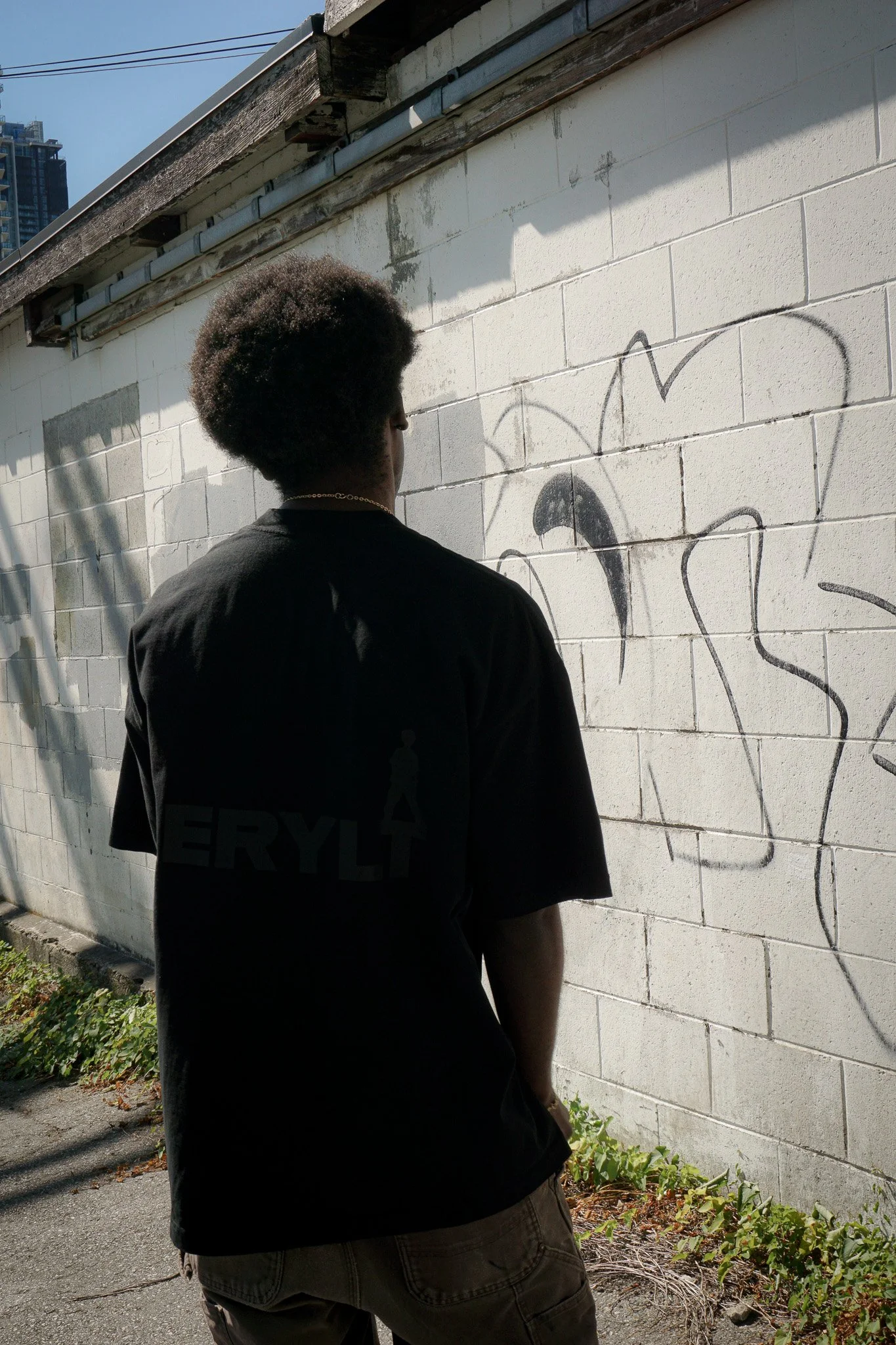 A person with an afro hairstyle wearing a black Aerylt T-shirt and shorts standing next to a white brick wall with graffiti. The person is facing the wall in a side profile, and buildings are visible in the background under a clear blue sky.
