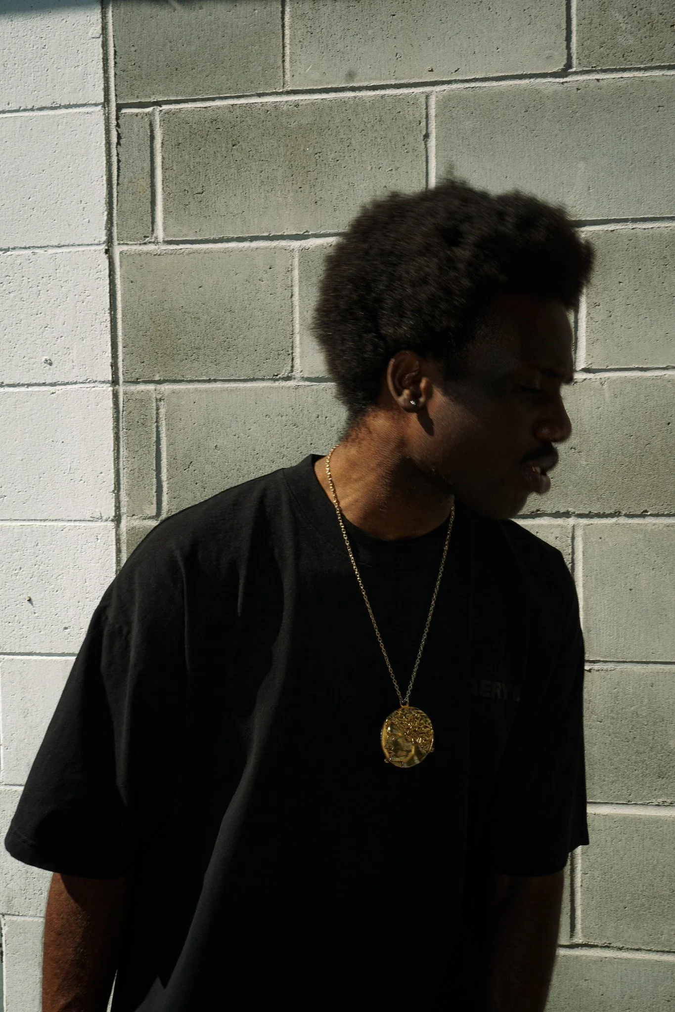 Side profile of a young Black woman with short curly hair, wearing a black Aerylt shirt, gold chain necklace with a large medallion, and earrings, standing against a cinder block wall.