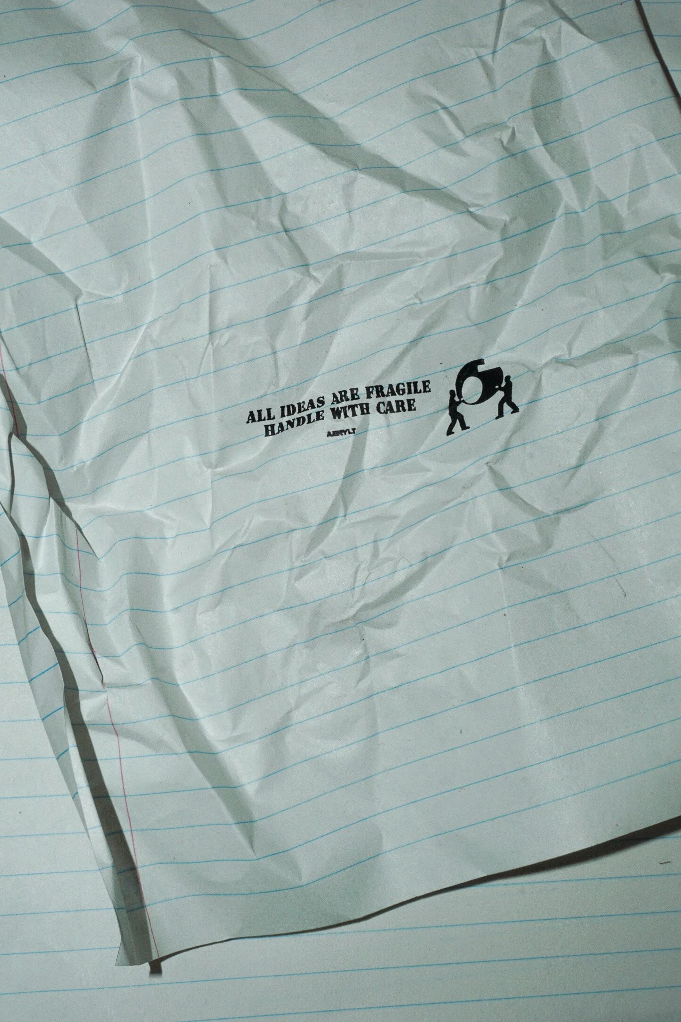 Crumpled sheet of white lined paper with a black and white sticker that reads, 'All ideas are fragile, handle with care. - Aerylt' accompanied by a small icon of the Aerylt Fragility Logo