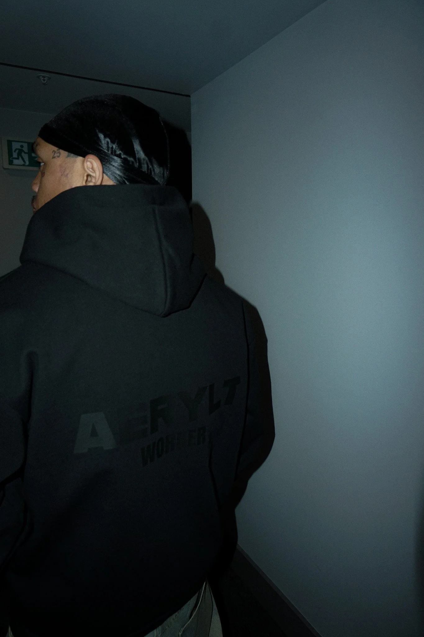 Person with a dark hat or durag, wearing a dark hoodie with Aerylt on the back, standing indoors near a white wall.