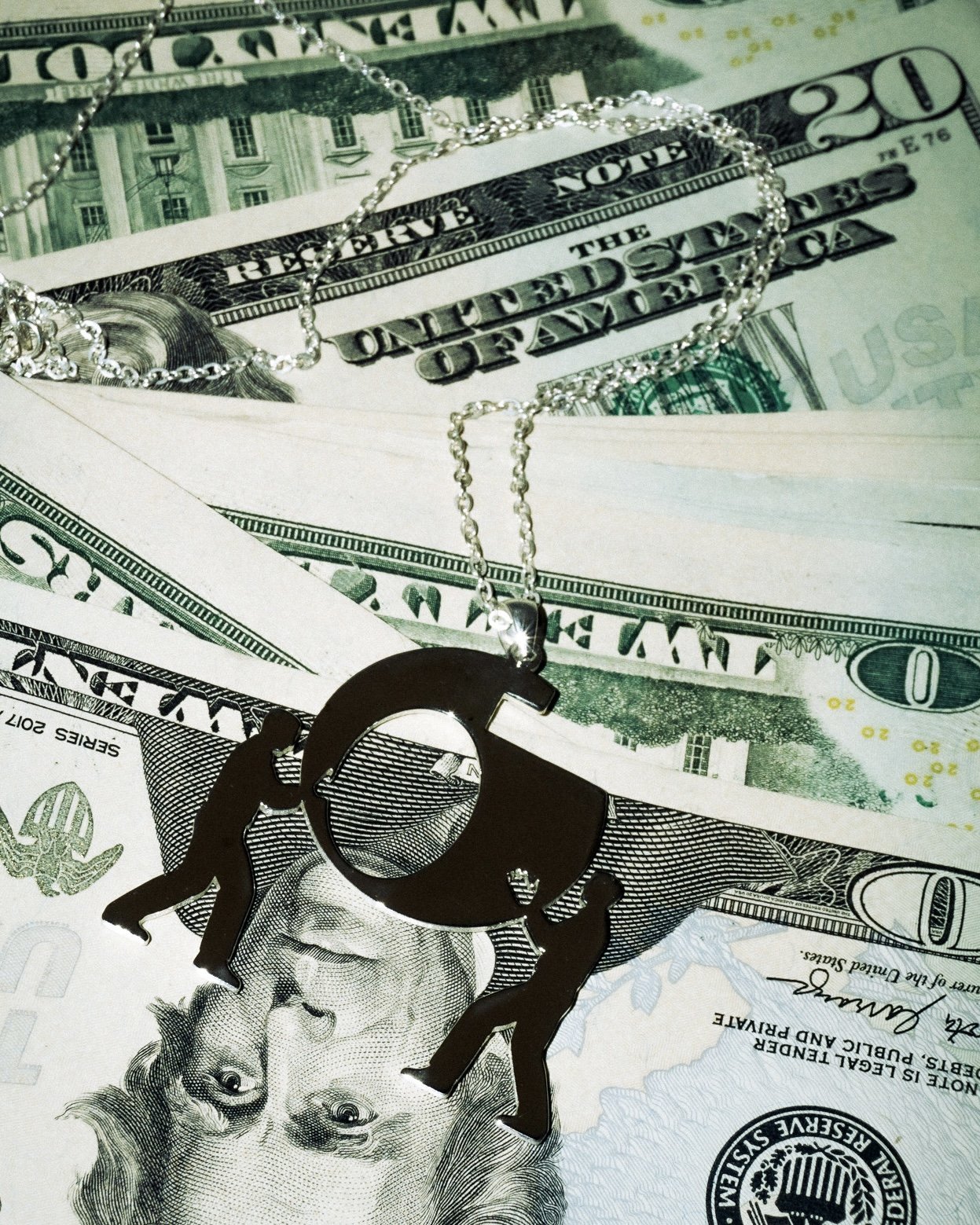 A silver necklace with a Aerylt Fragility Pendant top of various dollar bills, including a twenty-dollar bill and a one-hundred-dollar bill.
