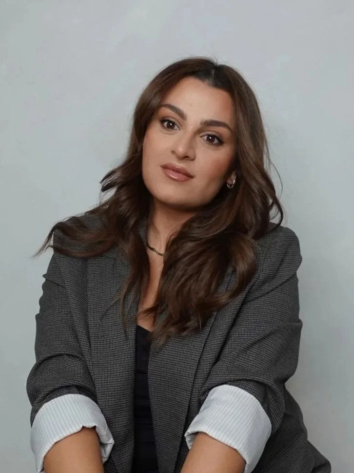 A young woman with wavy brown hair and neutral makeup, wearing a black top and a gray blazer with white cuffs, sitting against a plain light-colored wall.