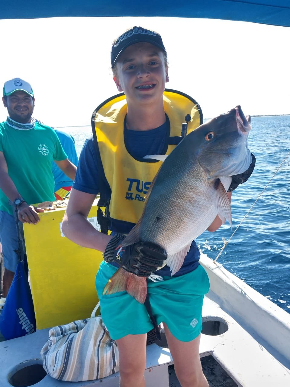 Teen boy holding a large fish on a boat, with two men in the background, on a sunny day at sea.