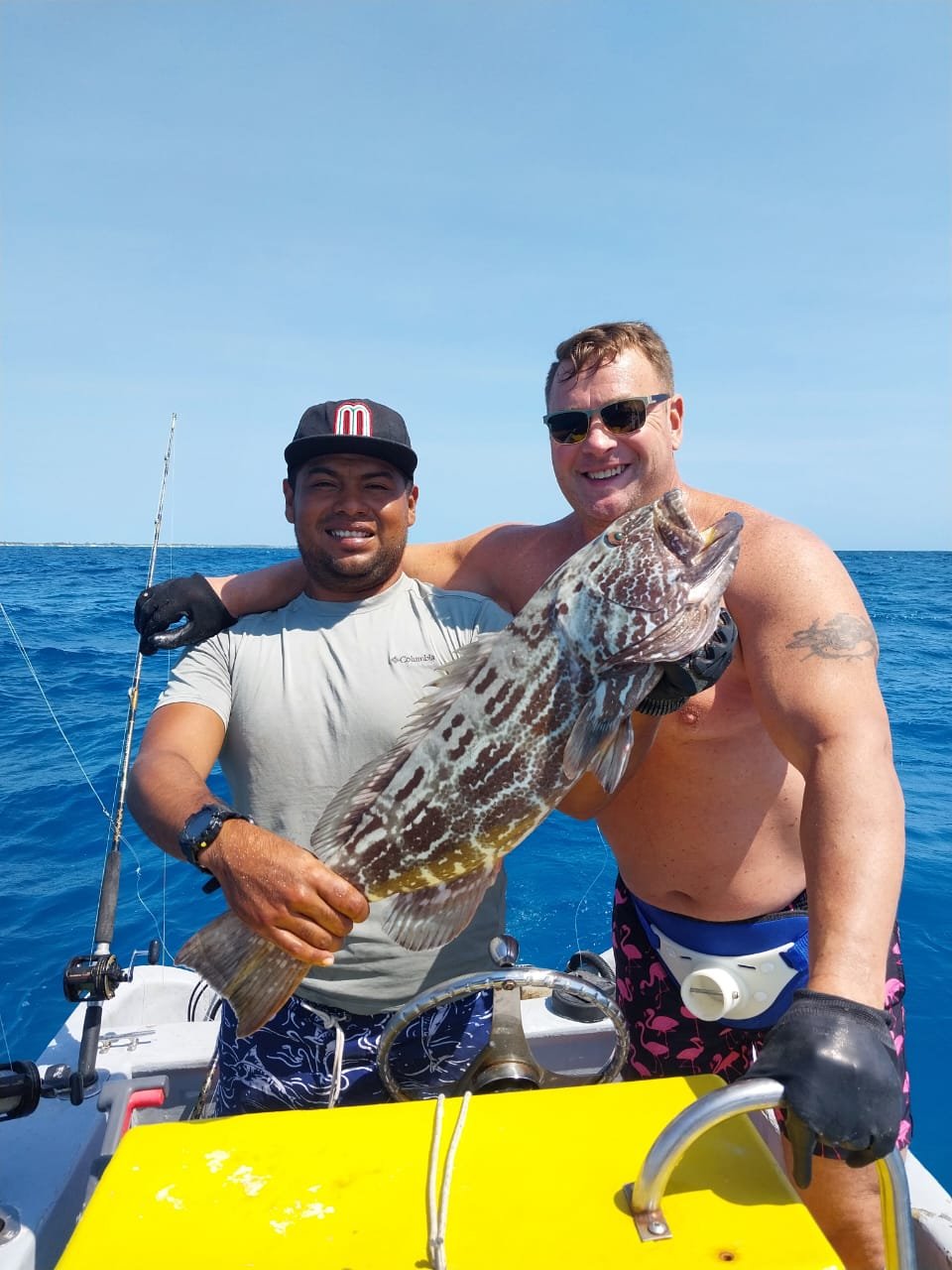 Two men on a boat holding a large fish, smiling, with the ocean in the background during daytime. One man is wearing a cap, and the other is shirtless with glasses.