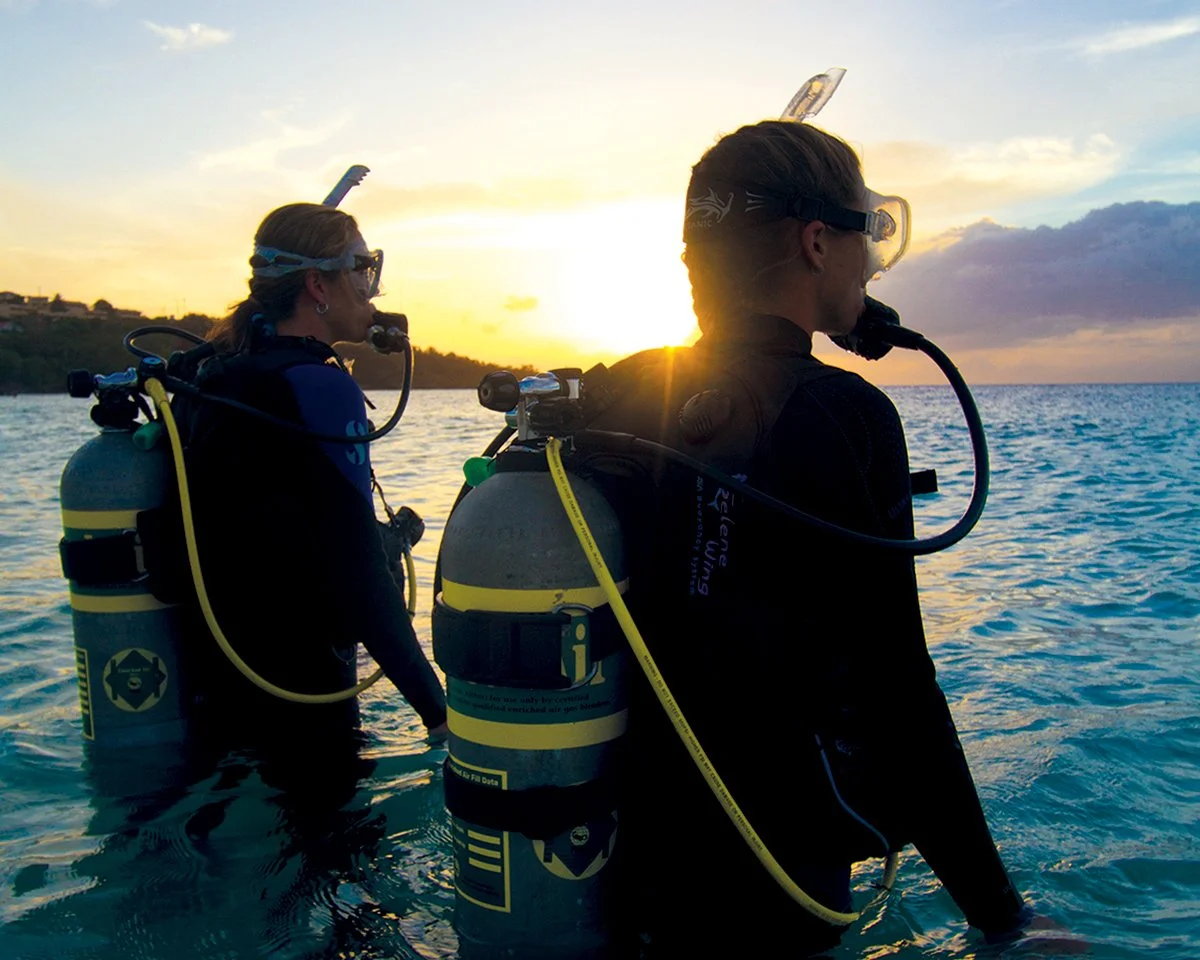 Two scuba divers in the ocean at sunset, preparing to dive with tanks and gear.