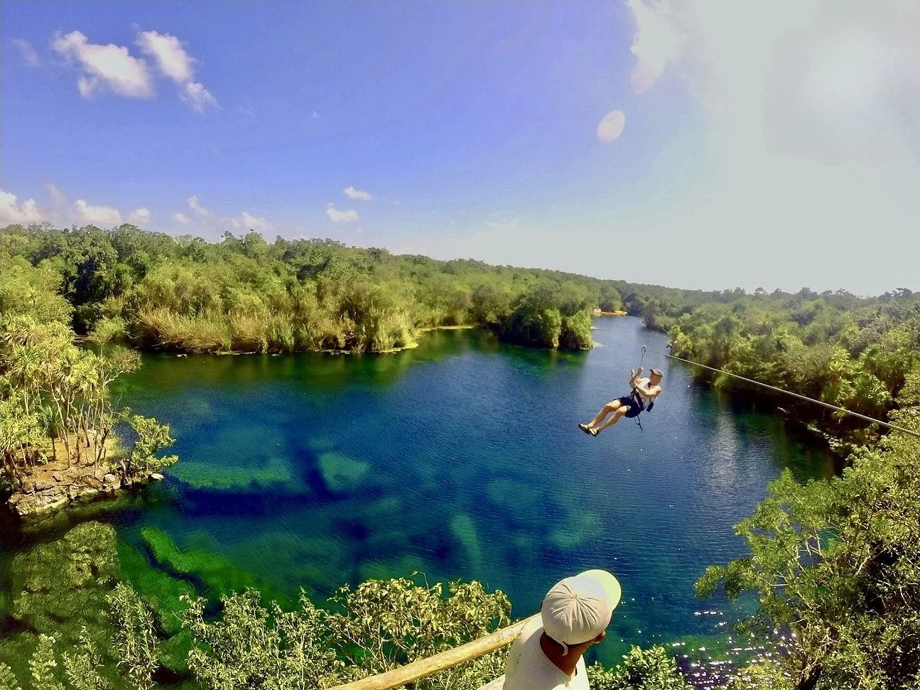 Person ziplining over a blue lake surrounded by lush green forest under a clear blue sky.