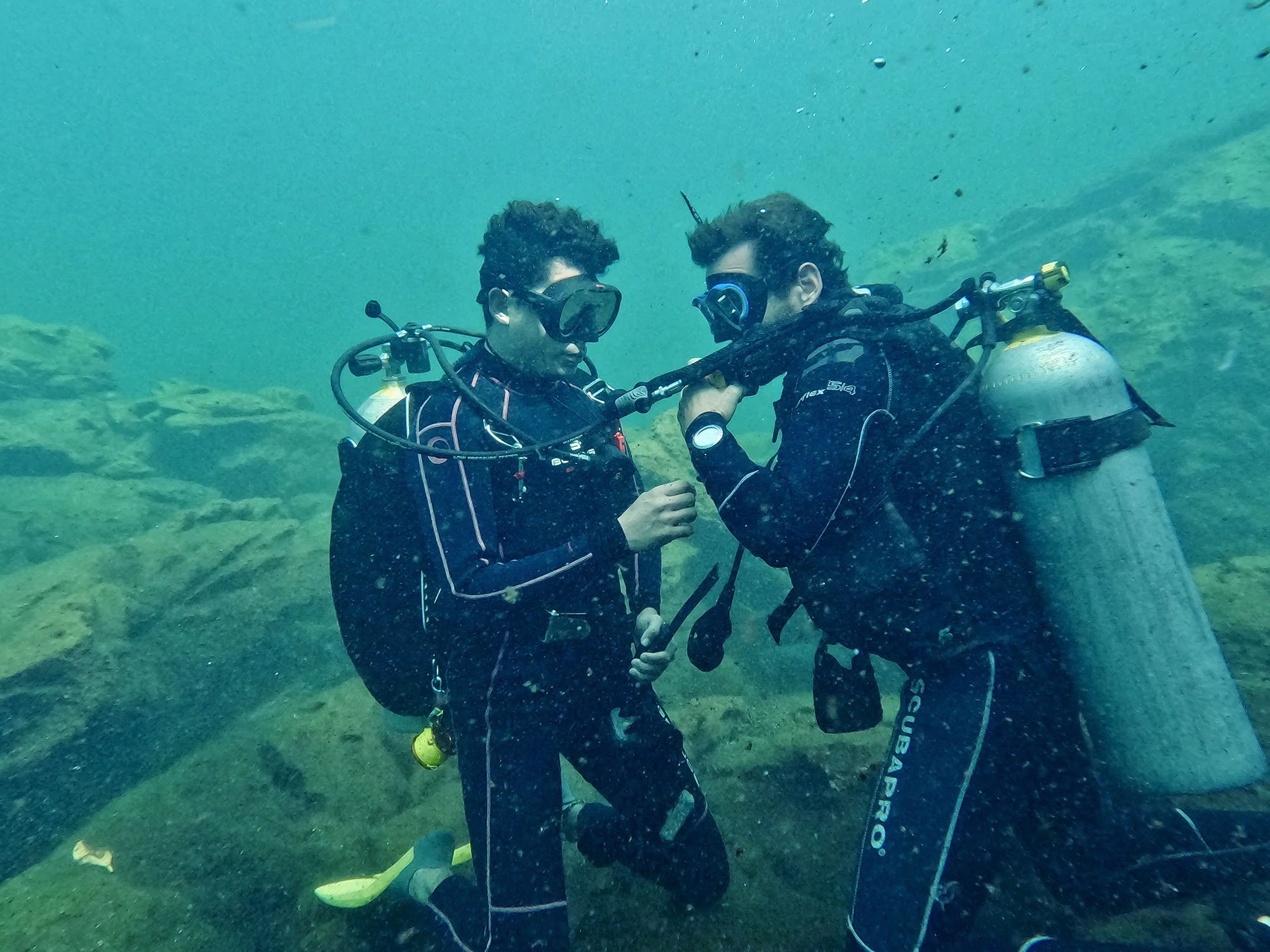 Two scuba divers underwater in wetsuits and masks, one holding a diving regulator, positioned over a rocky seabed.