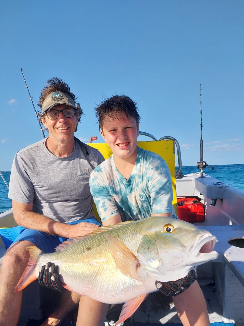 A man and a boy sitting on a boat, smiling, with the boy holding a large fish they caught, under a clear blue sky.