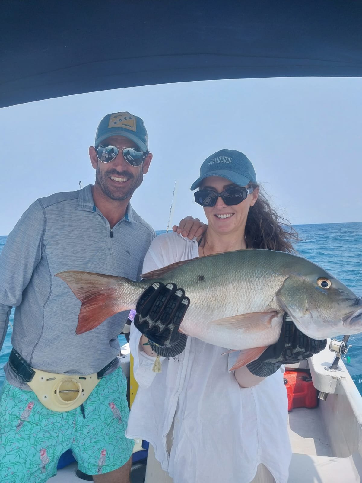 A man and woman on a boat holding a large fish they caught while fishing at sea. The woman is holding the fish with both hands, and both are smiling at the camera.