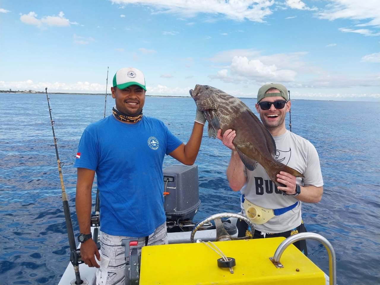 Two men on a boat holding a large fish, with fishing rods and the ocean in the background, smiling and enjoying their catch.