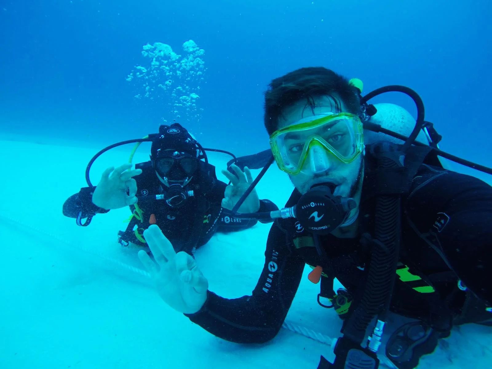 Two scuba divers underwater making OK hand signals and wearing diving gear.