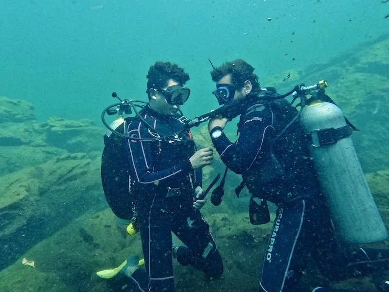 Two scuba divers underwater in wetsuits and masks, one holding a diving regulator, positioned over a rocky cenote bed, in a rescue diver course
