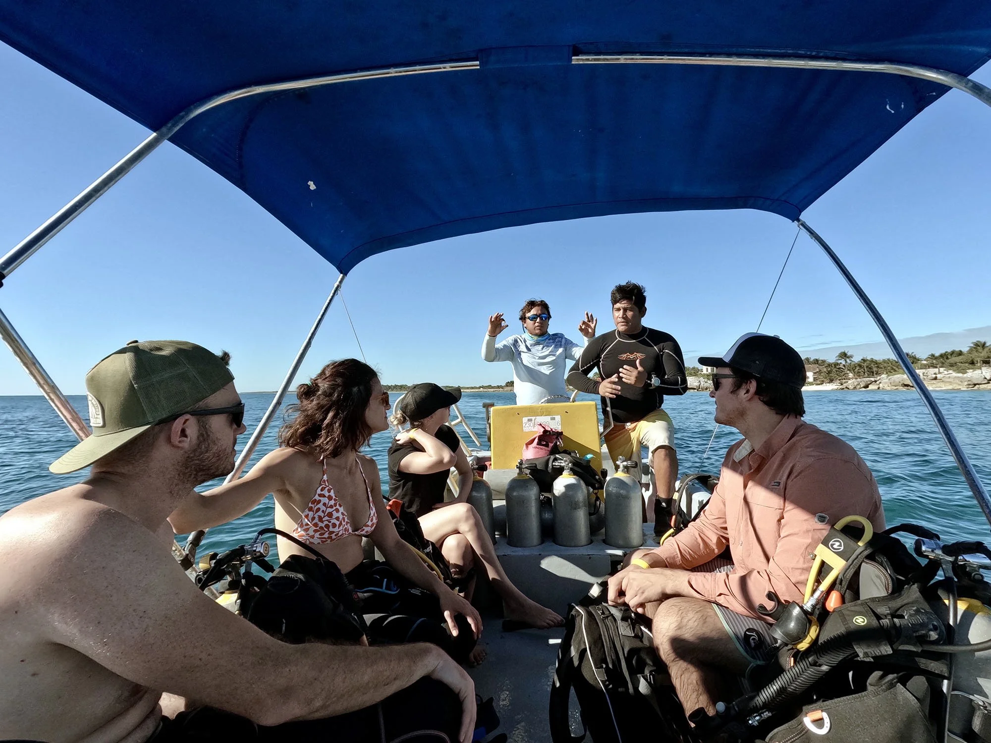 Group of people sitting in a small boat with scuba diving gear, under a blue canopy, on a sunny day at sea.