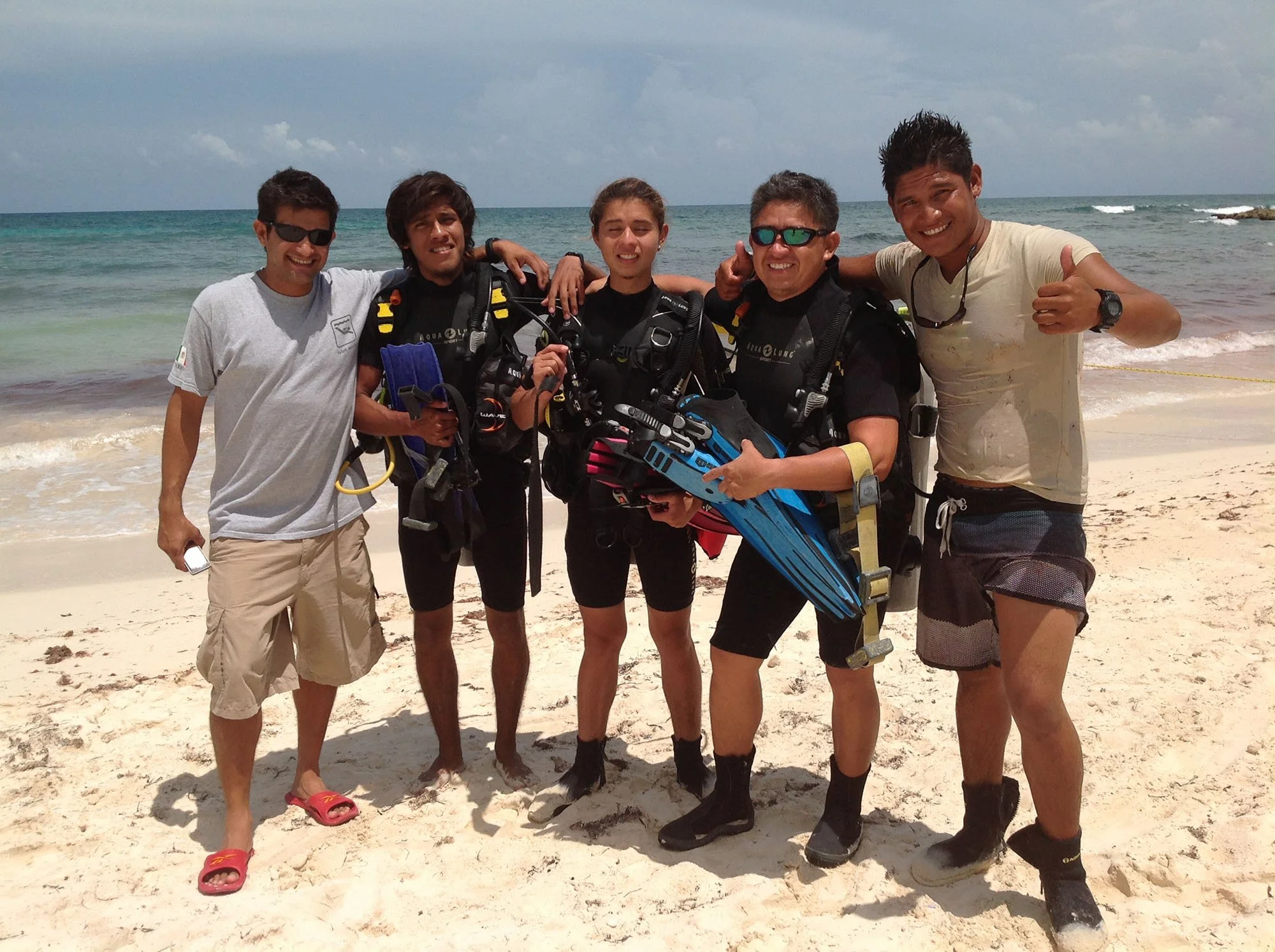 Group of five men standing on a beach, holding scuba diving gear, with ocean and sky in the background.