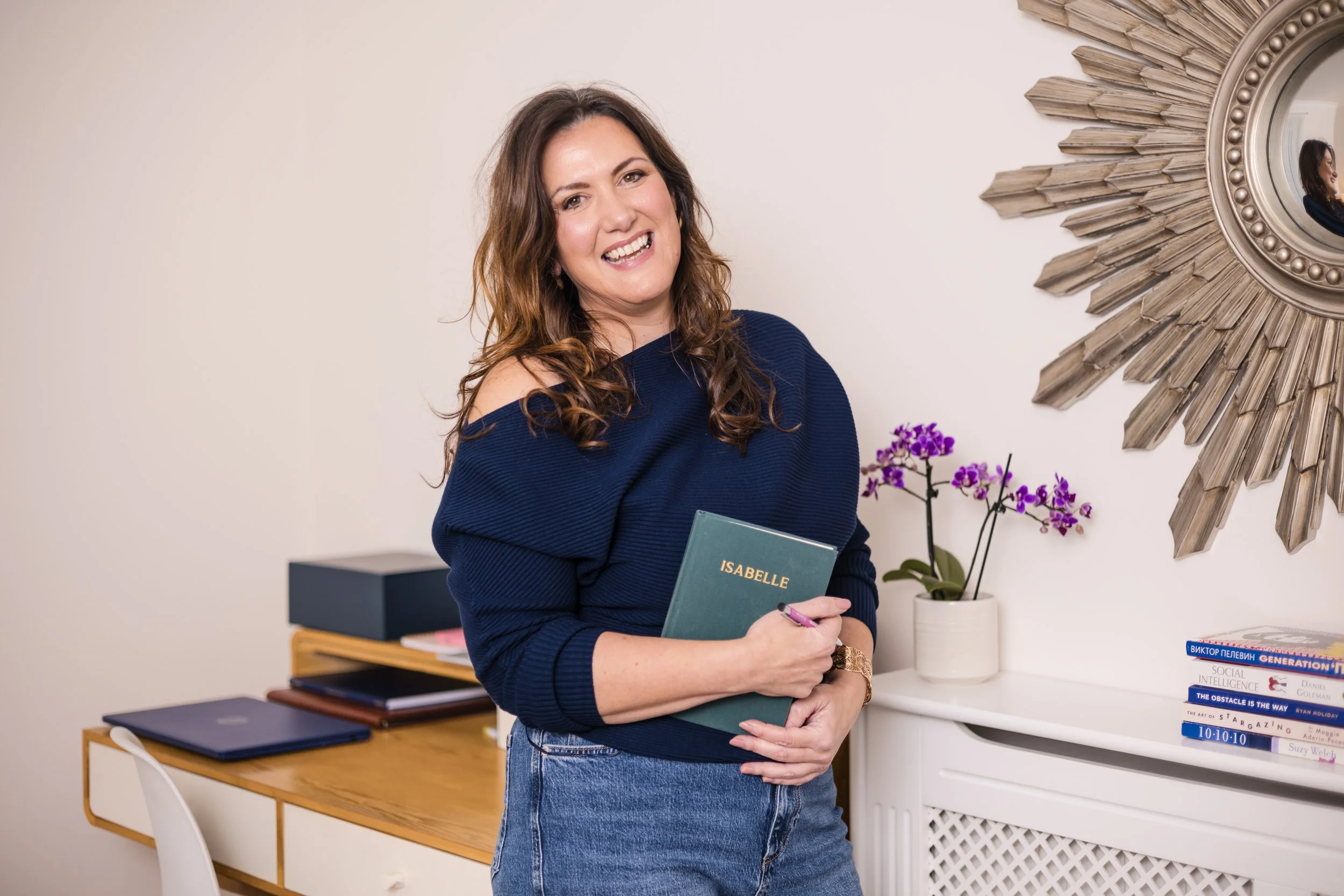 A woman with brown hair, wearing a navy blue sweater and jeans, smiling and holding a green notebook labeled 'ISABELLE' in an indoor setting with a desk, a stack of books, and a decorative sunburst mirror on the wall.