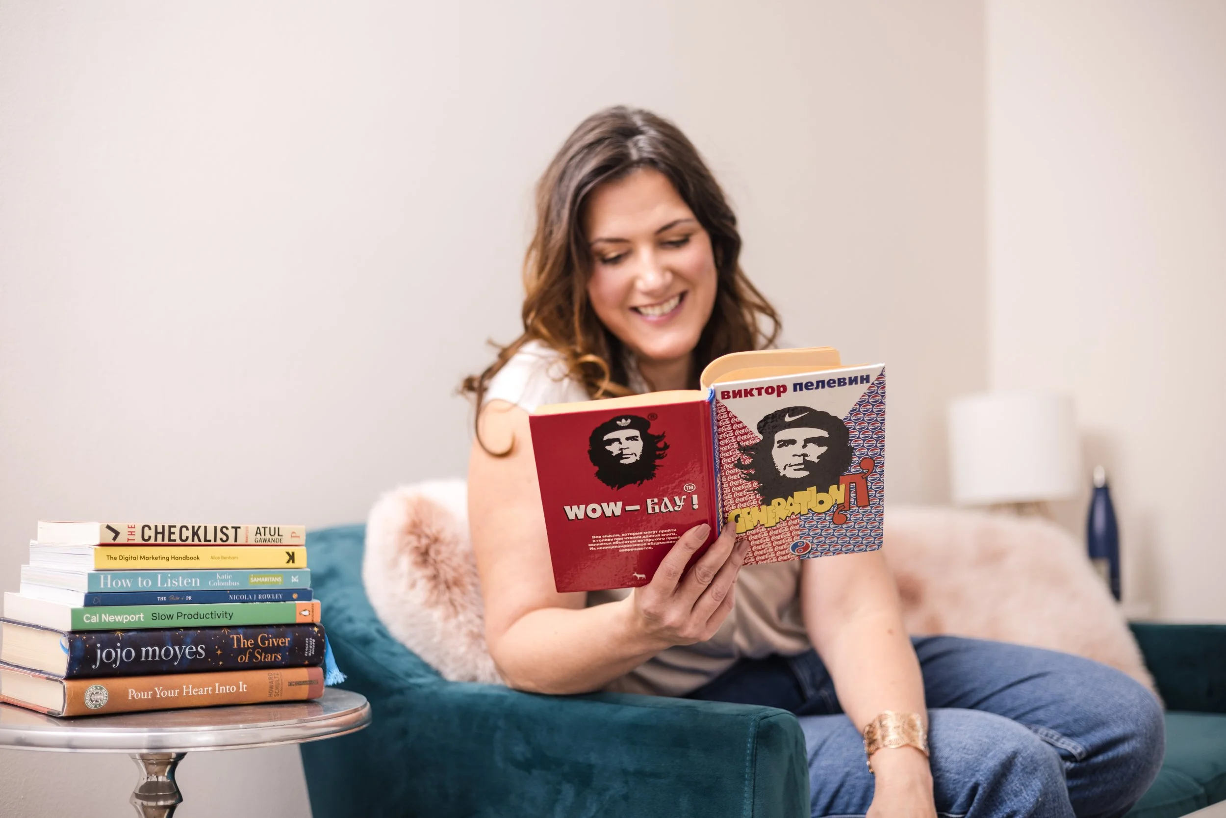 A woman sitting on a teal soft velvet sofa, smiling and reading a Russian book. Beside her, a stack of books is placed on a glass side table. The room has a neutral-colored wall and a white lamp in the background.