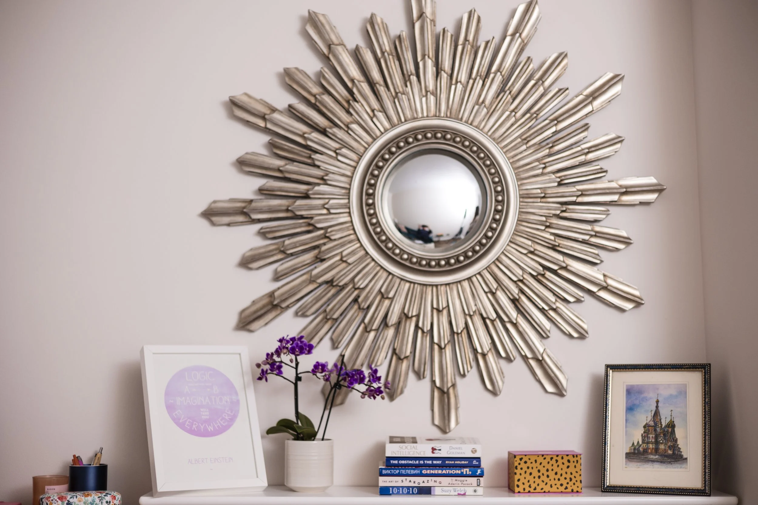 Decorative wall mirror with a sunburst design hanging above a white shelf. On the shelf are framed art, a potted purple orchid, a stack of books, a yellow polka dot box, and a framed painting of a castle.