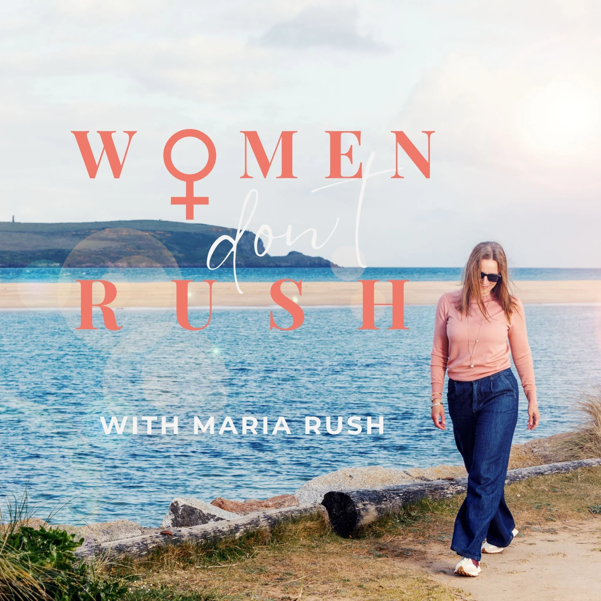 Woman walking along a rocky shoreline near the ocean, with hills in the background, wearing sunglasses, a pink jacket, and jeans.