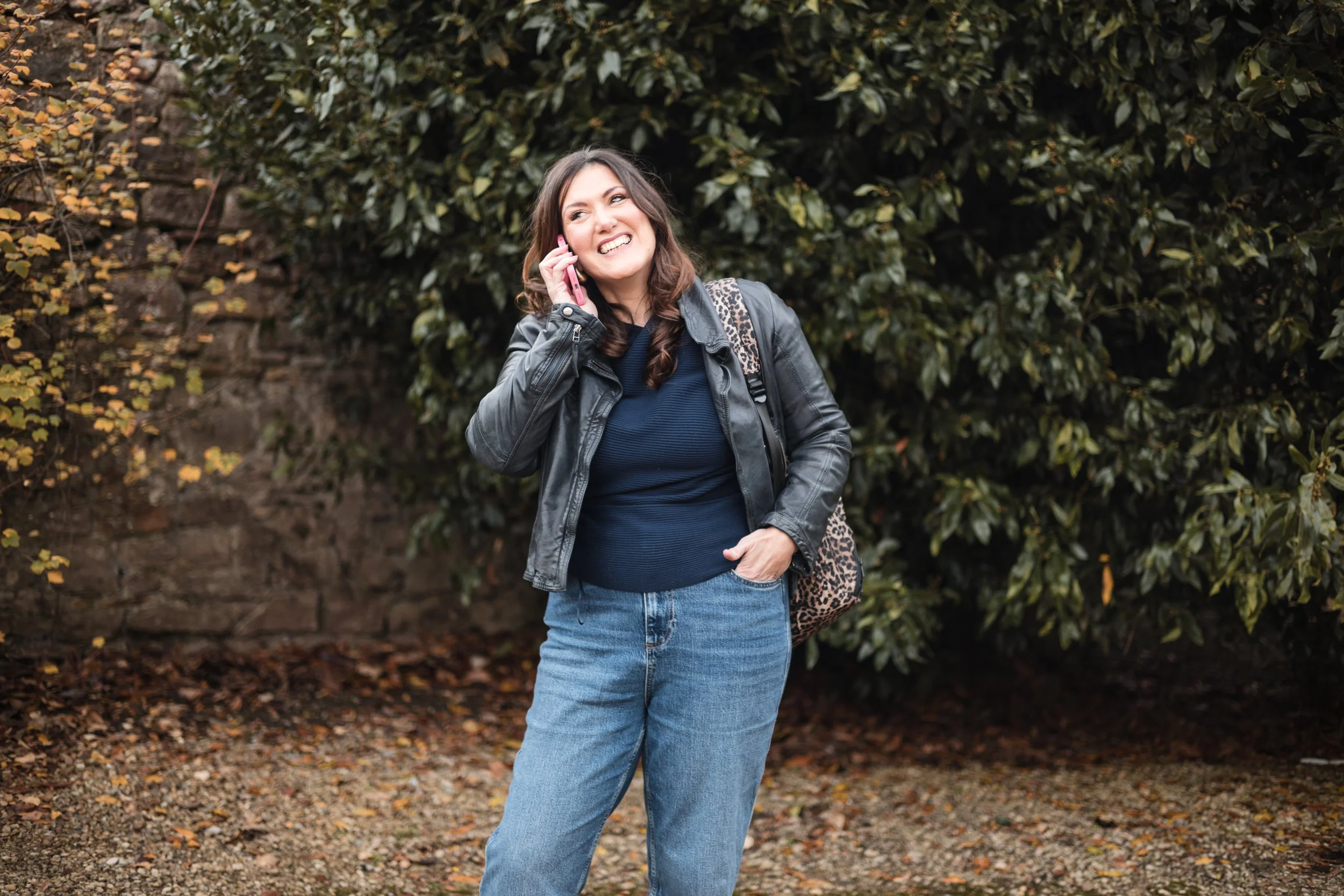 A woman with brown hair talking on her smartphone outdoors, wearing a navy short leather jacket, a blue top, jeans, and a backpack with a leopard print pattern, standing in front of large green bushes.