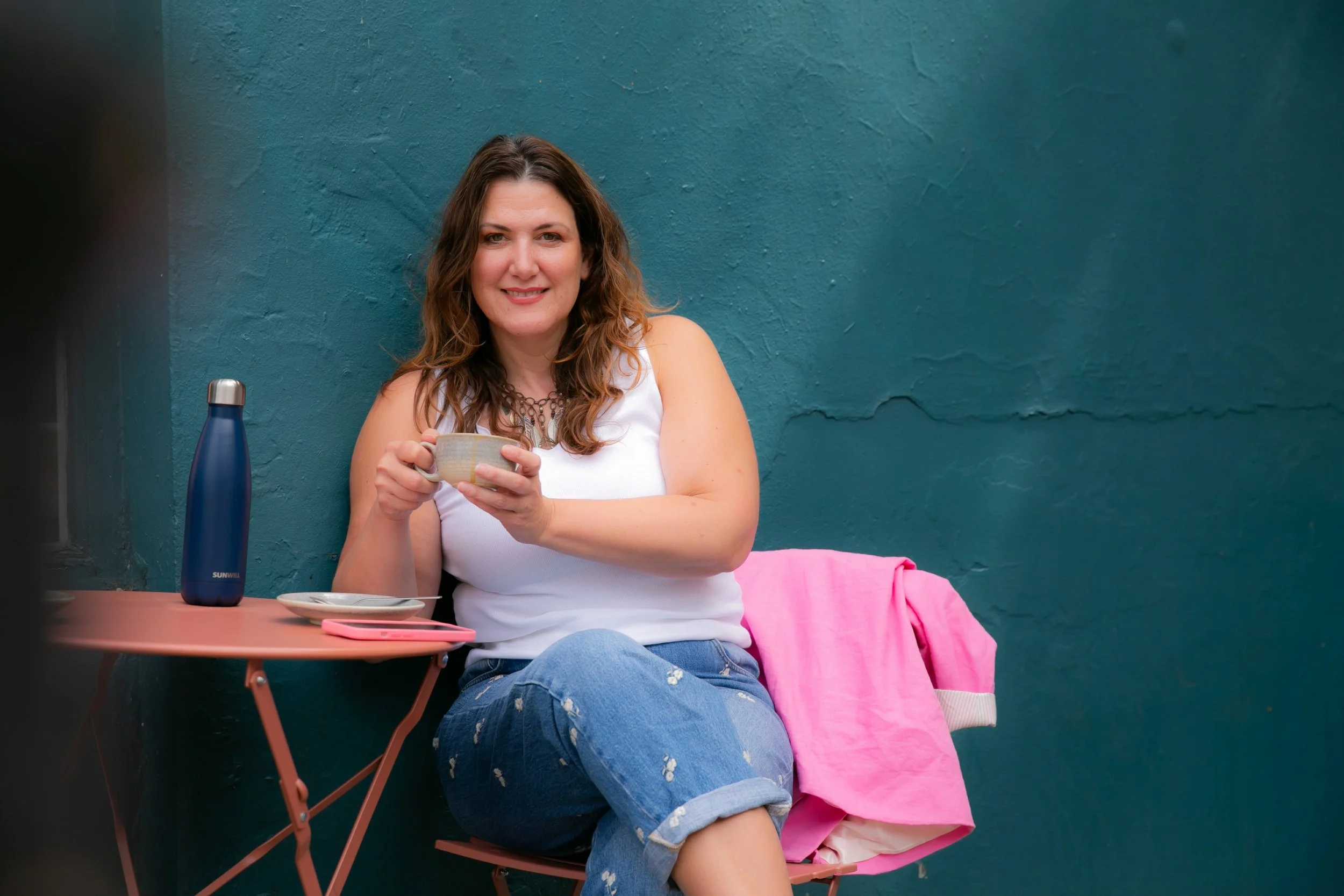 A woman with brown wavy hair and fair skin sitting outdoors at a pink table, drinking from a beige cup, with a teal wall behind her. She is wearing a white sleeveless top and ripped blue jeans, and has a pink blanket or jacket draped over her lap. There is a blue water bottle and a pink phone on the table.