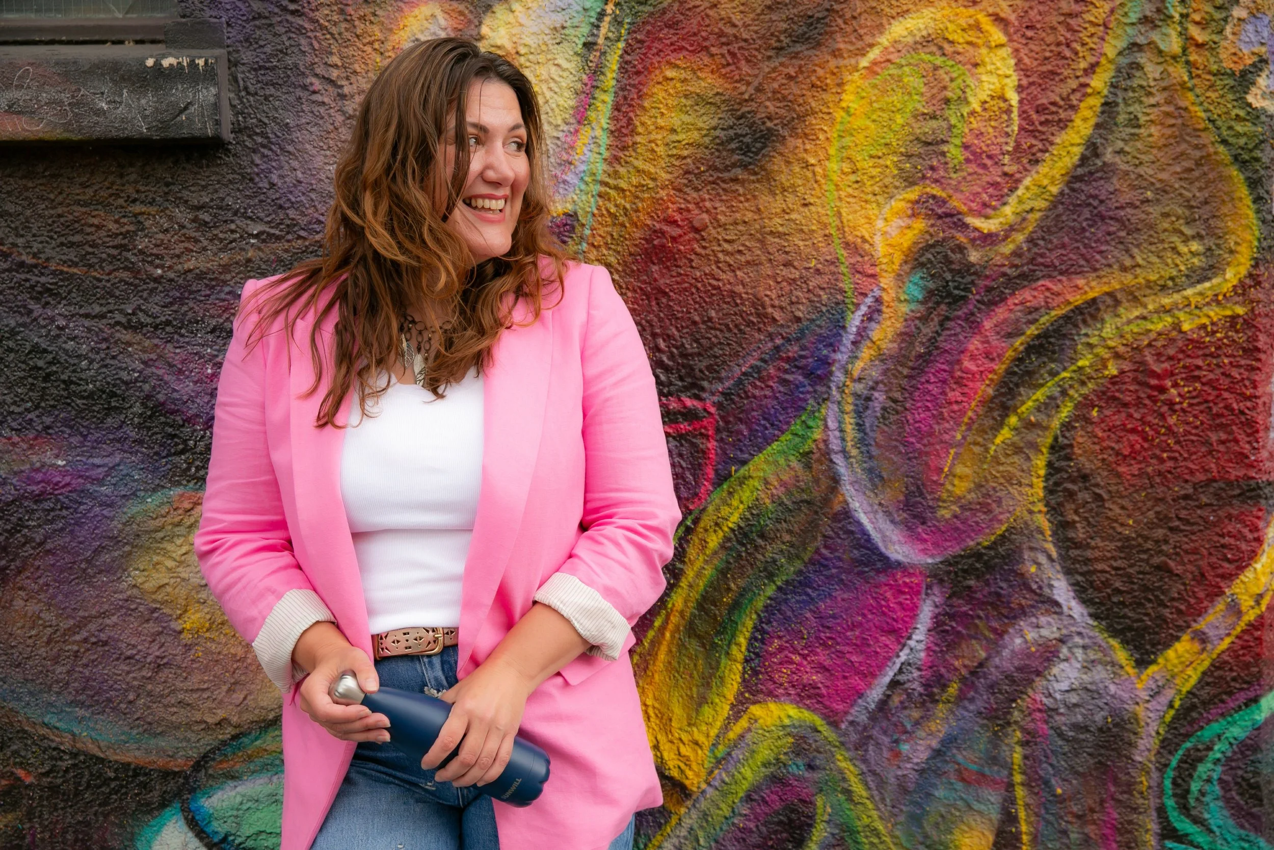 Woman with wavy brown hair in a pink blazer and white top smiling and holding a spray bottle, standing against a colorful, textured graffiti wall.