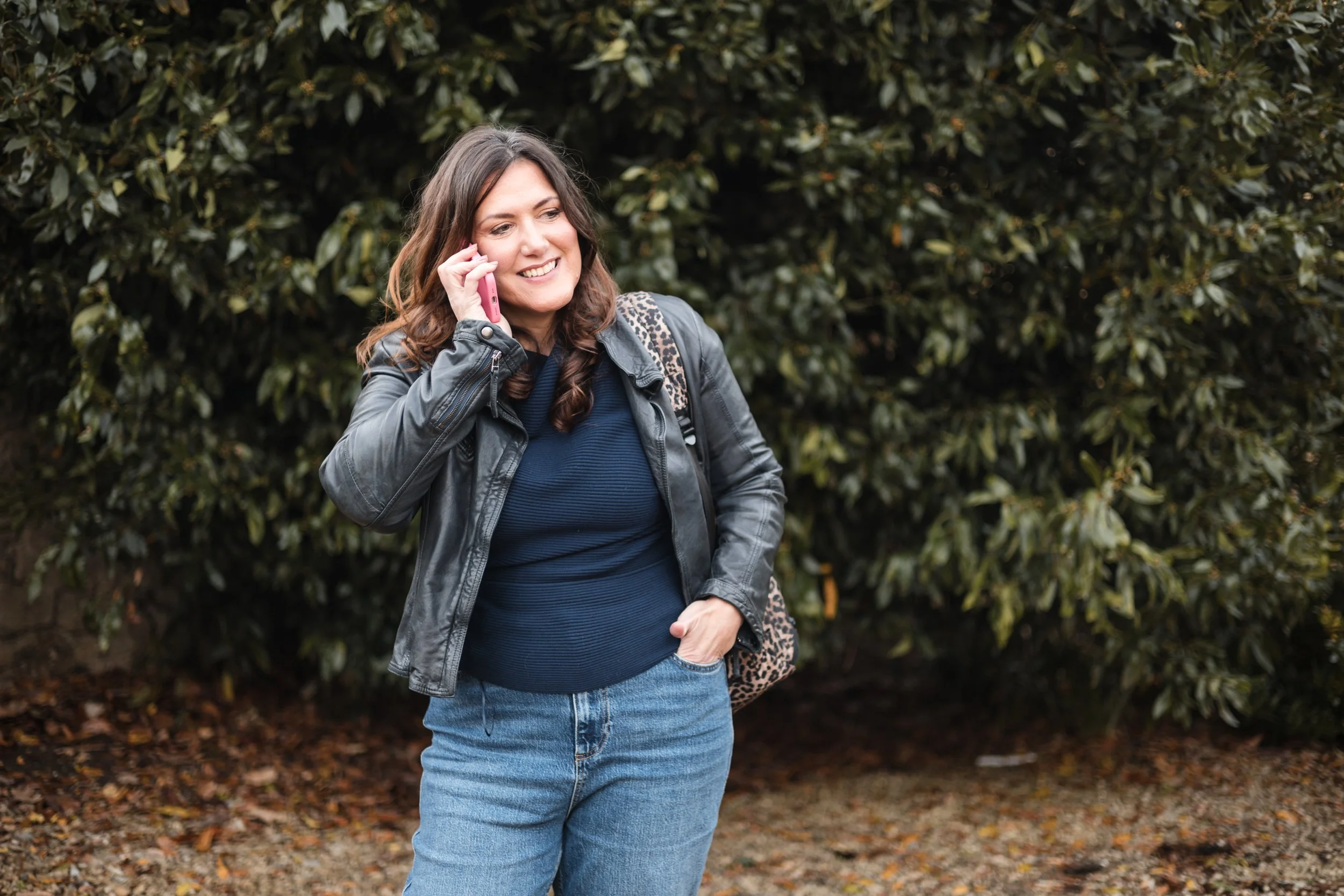 A woman with brown hair, wearing a navy leather jacket, navy blue top, and blue jeans, talking on a pink smartphone outdoors near a large green bush, smiling and holding her hand near her jeans pocket.