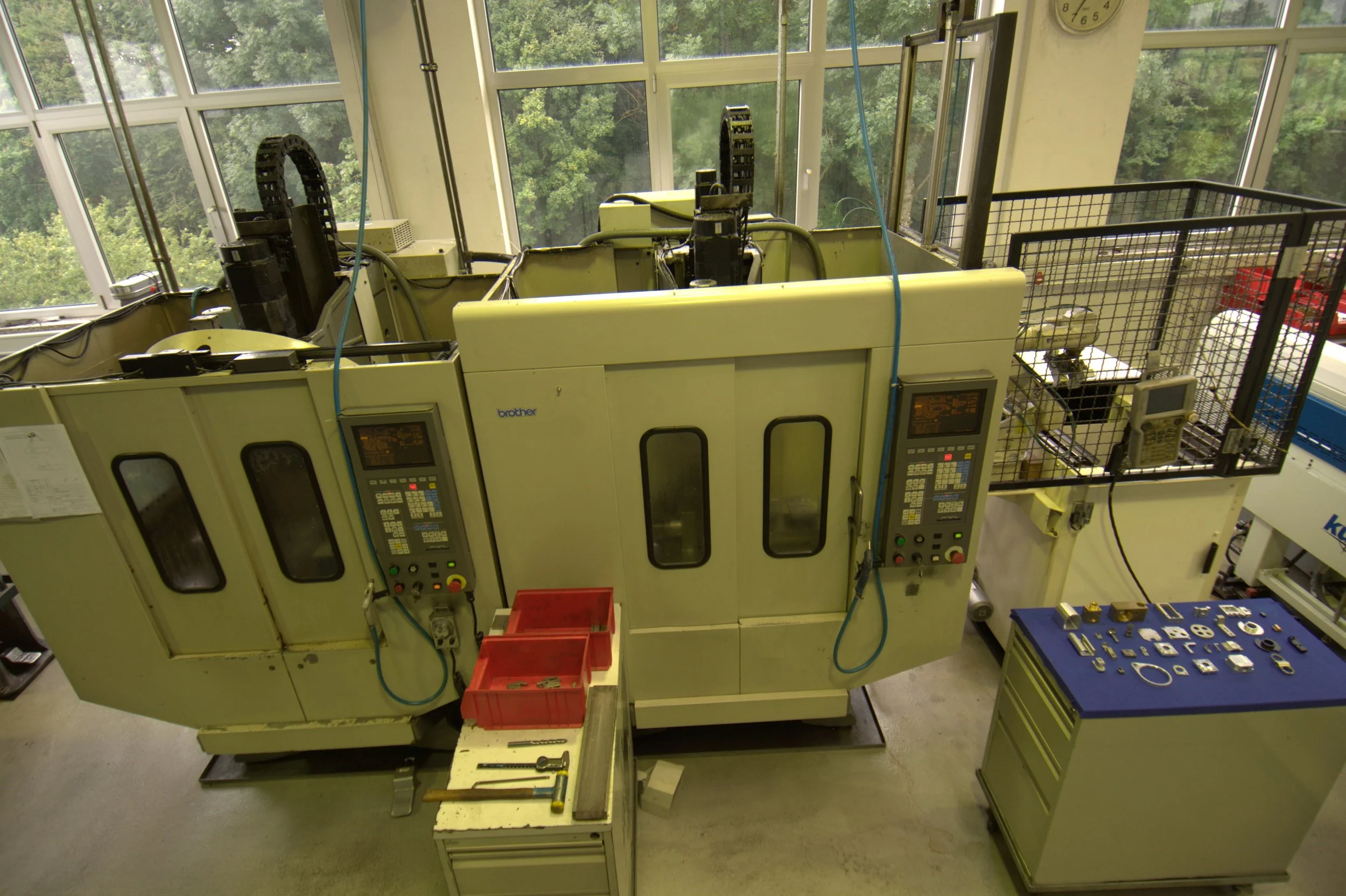 Hochpräzise Fertigungsmaschine in einer Werkshalle mit großen Fenstern, Werkzeugen auf einem Tisch und technischen Komponenten auf einem blauen Tisch.