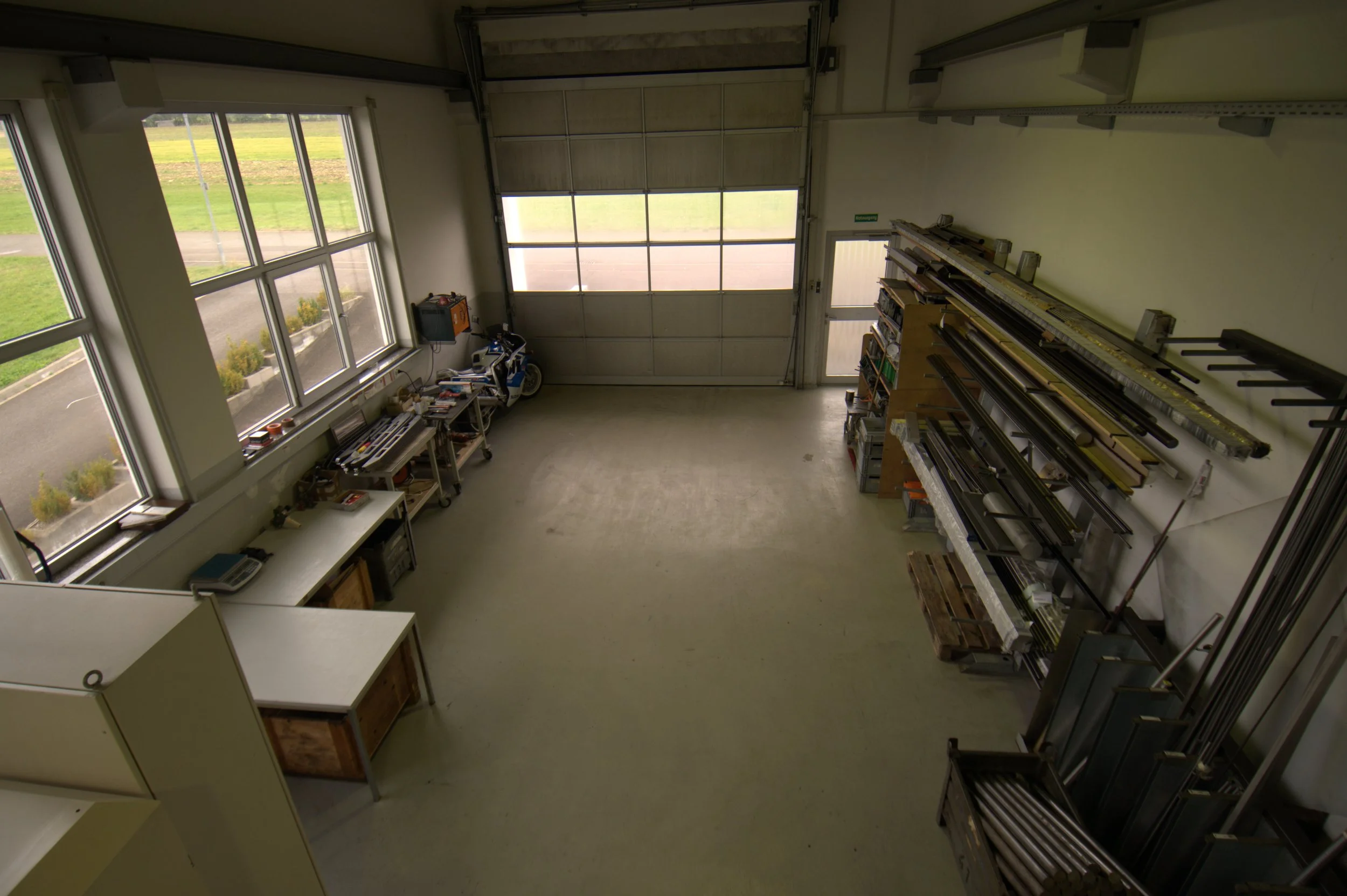 Innenansicht einer Werkstatt mit großen Fenstern, einem Schreibtisch, Werkzeugen, Regalen und Garagentor